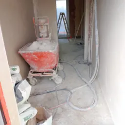 Wnętrze w trakcie prac tynkarskich: maszyna do tynkowania, worki z zaprawą, świeżo otynkowane ściany i drabina w tle. Widoczne przewody i węże.