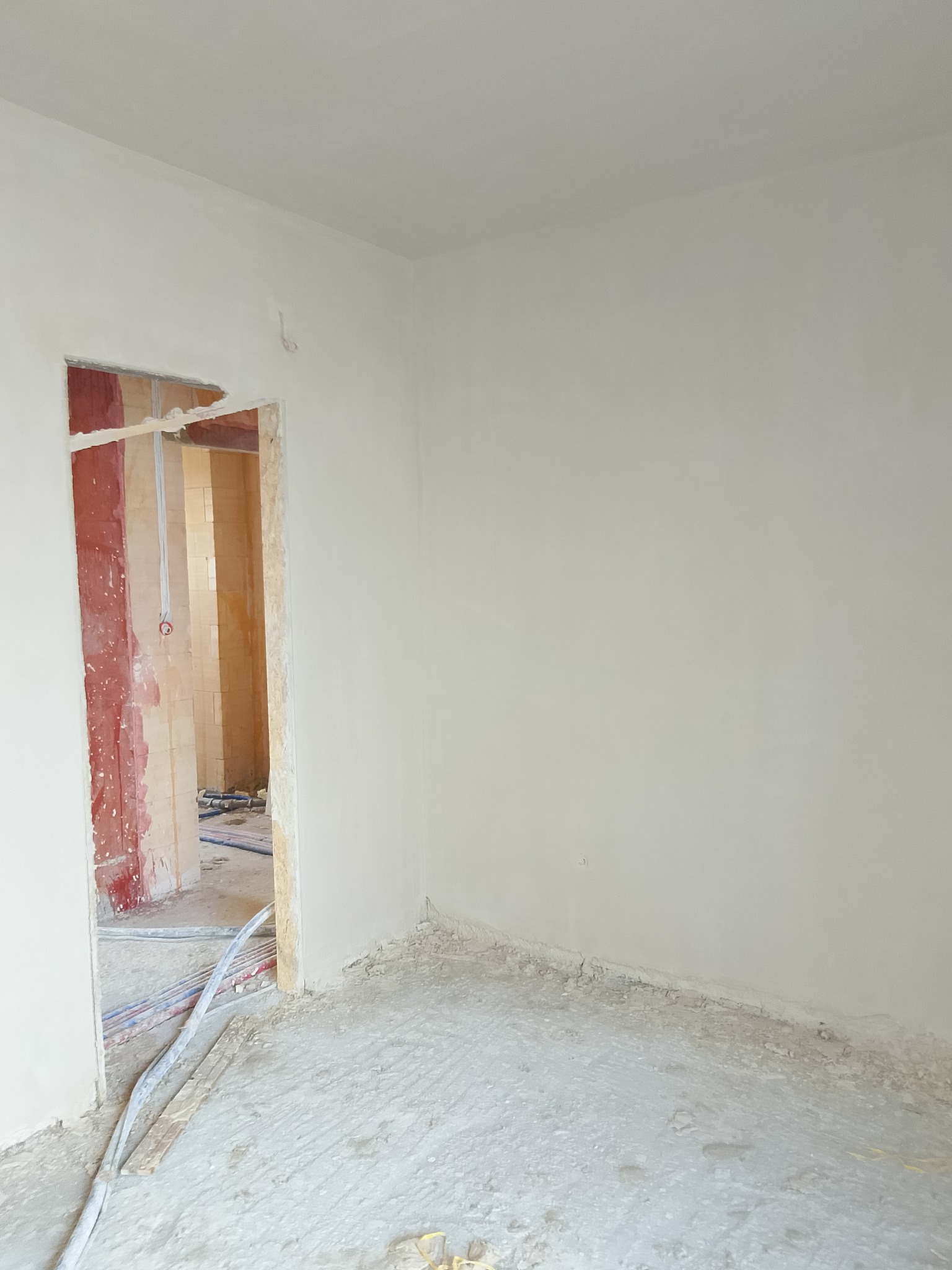 Wnętrze po tynkowaniu: gładkie, jasne ściany i sufit. Widoczne przewody i instalacje w przejściu do sąsiedniego pomieszczenia, przygotowane do dalszych prac wykończeniowych.
