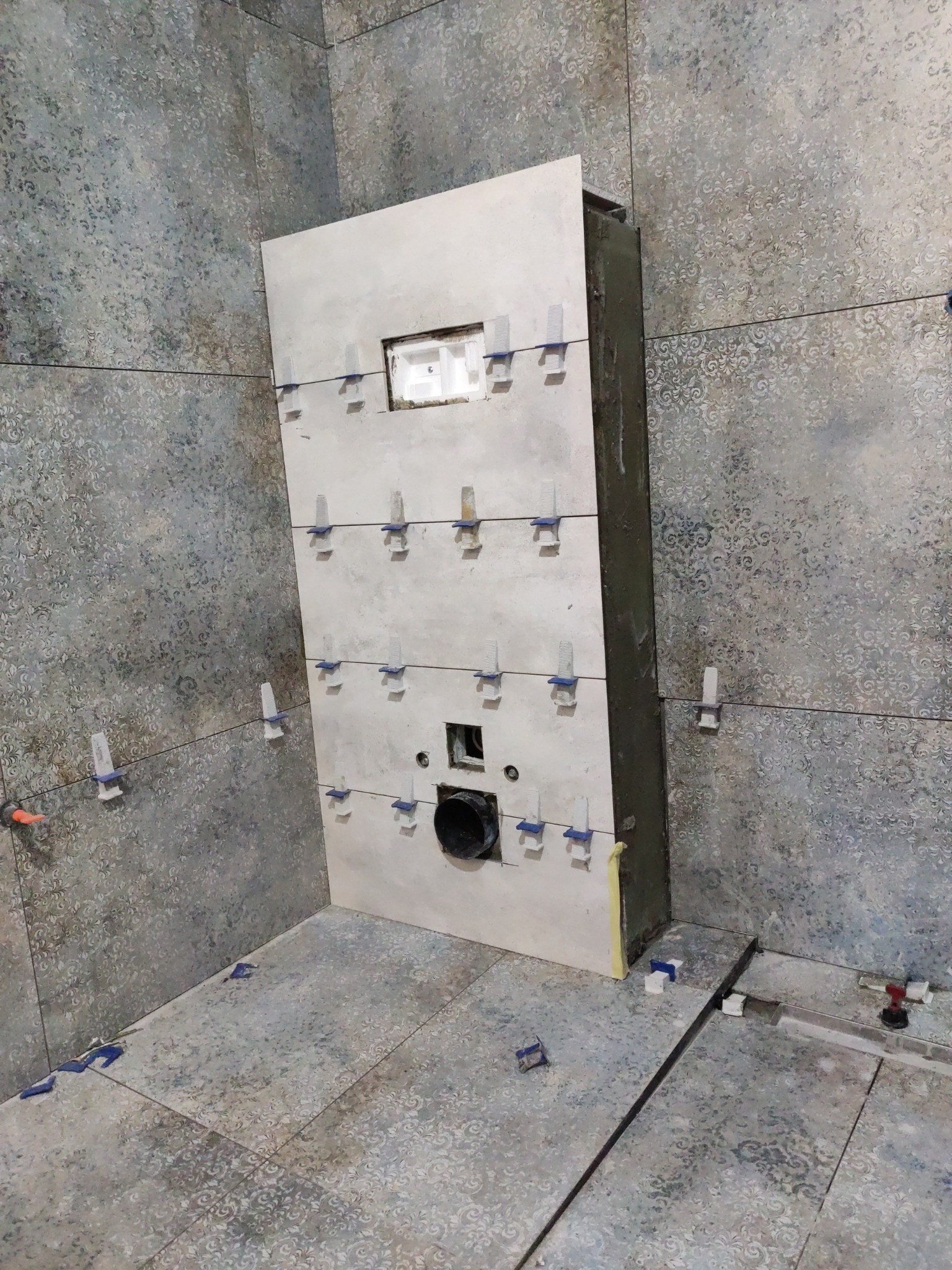 Ściana w łazience z płytkami w trakcie montażu, widoczne krzyżyki dystansowe. Fragment pod zabudowę stelaża WC z otworem na rurę kanalizacyjną.