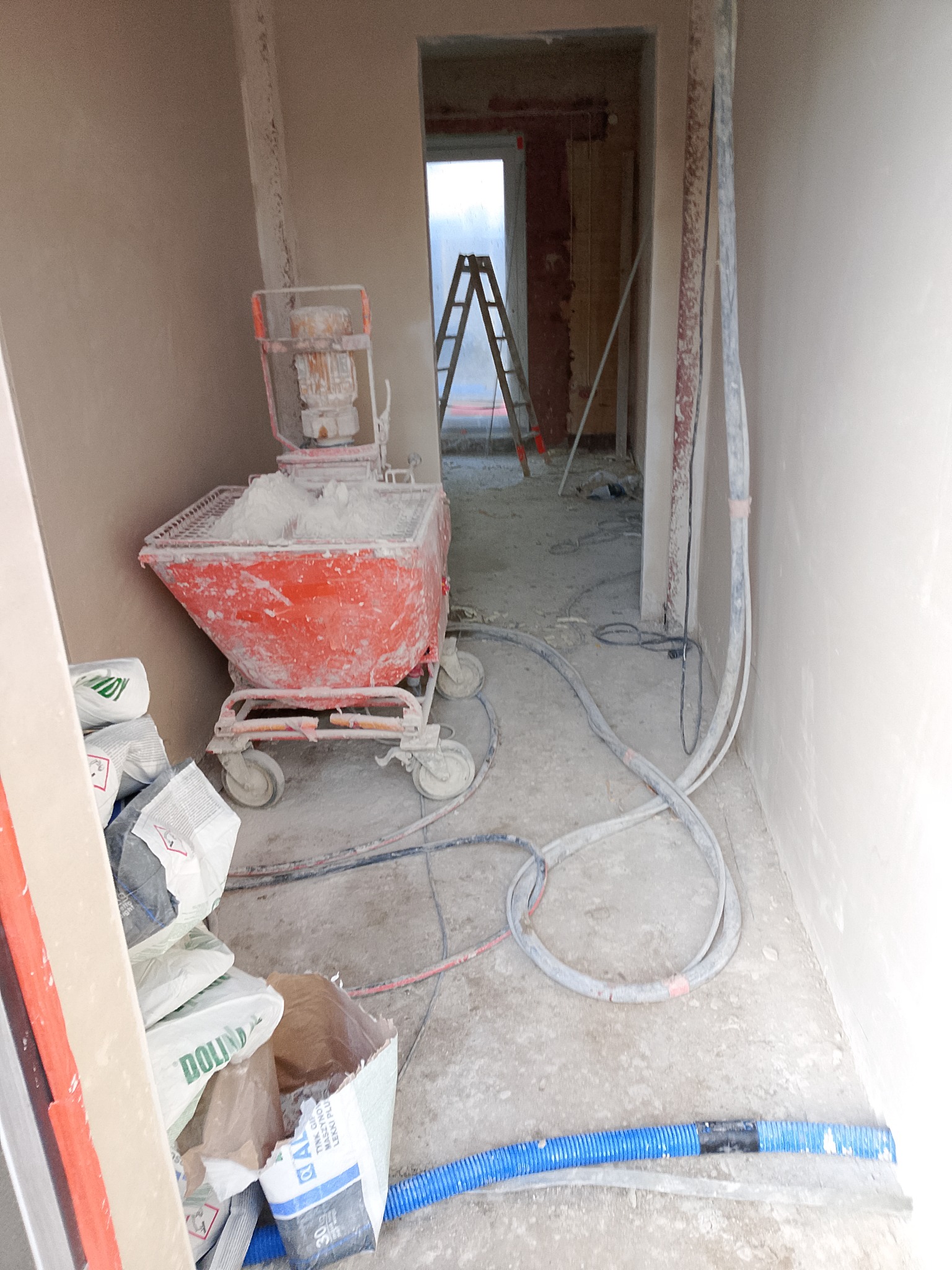 Wnętrze w trakcie prac tynkarskich: maszyna do tynkowania, worki z zaprawą, świeżo otynkowane ściany i drabina w tle. Widoczne przewody i węże.