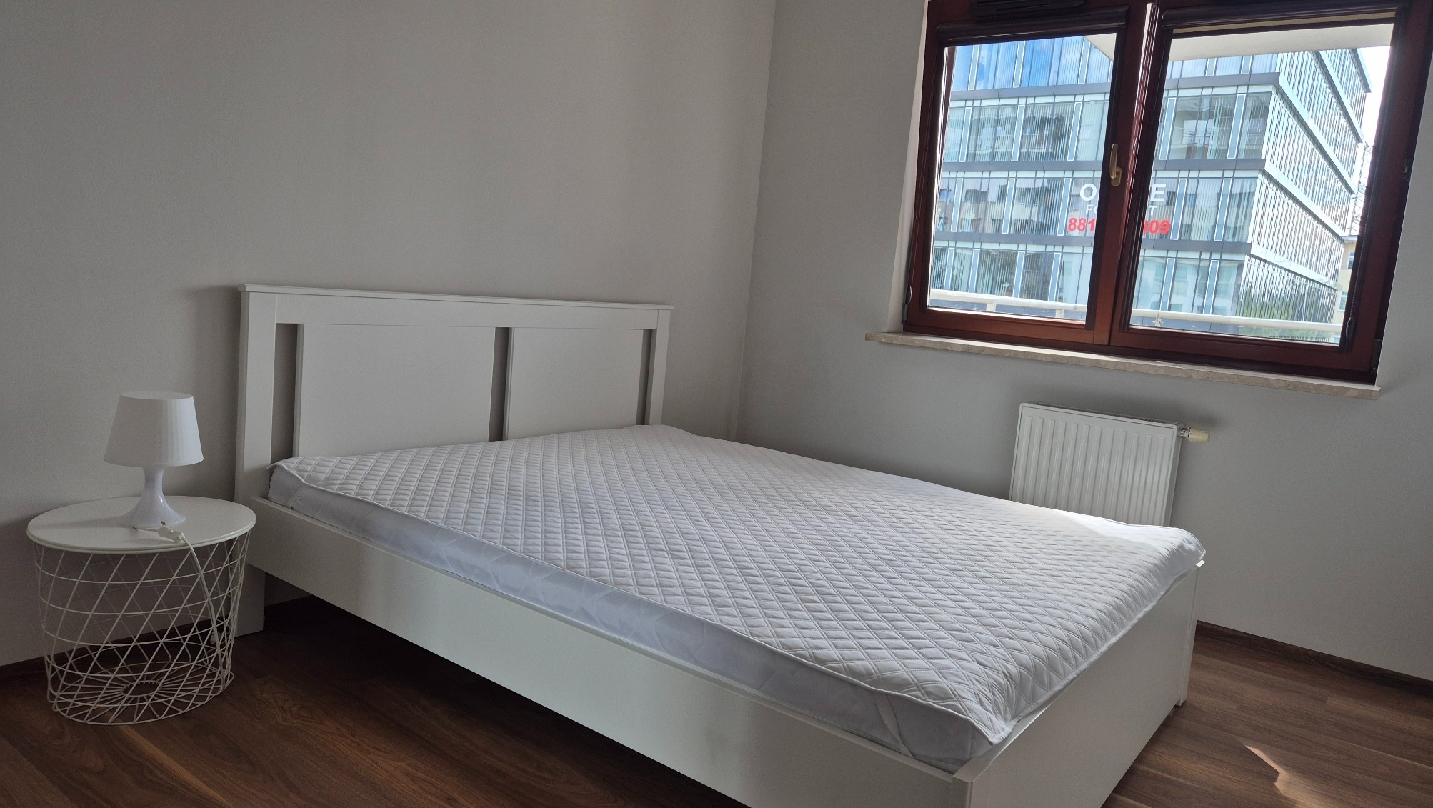 Jasna sypialnia z białym łóżkiem i stolikiem nocnym. Za oknem widoczny nowoczesny budynek biurowy. Minimalistyczny wystrój, drewniana podłoga.