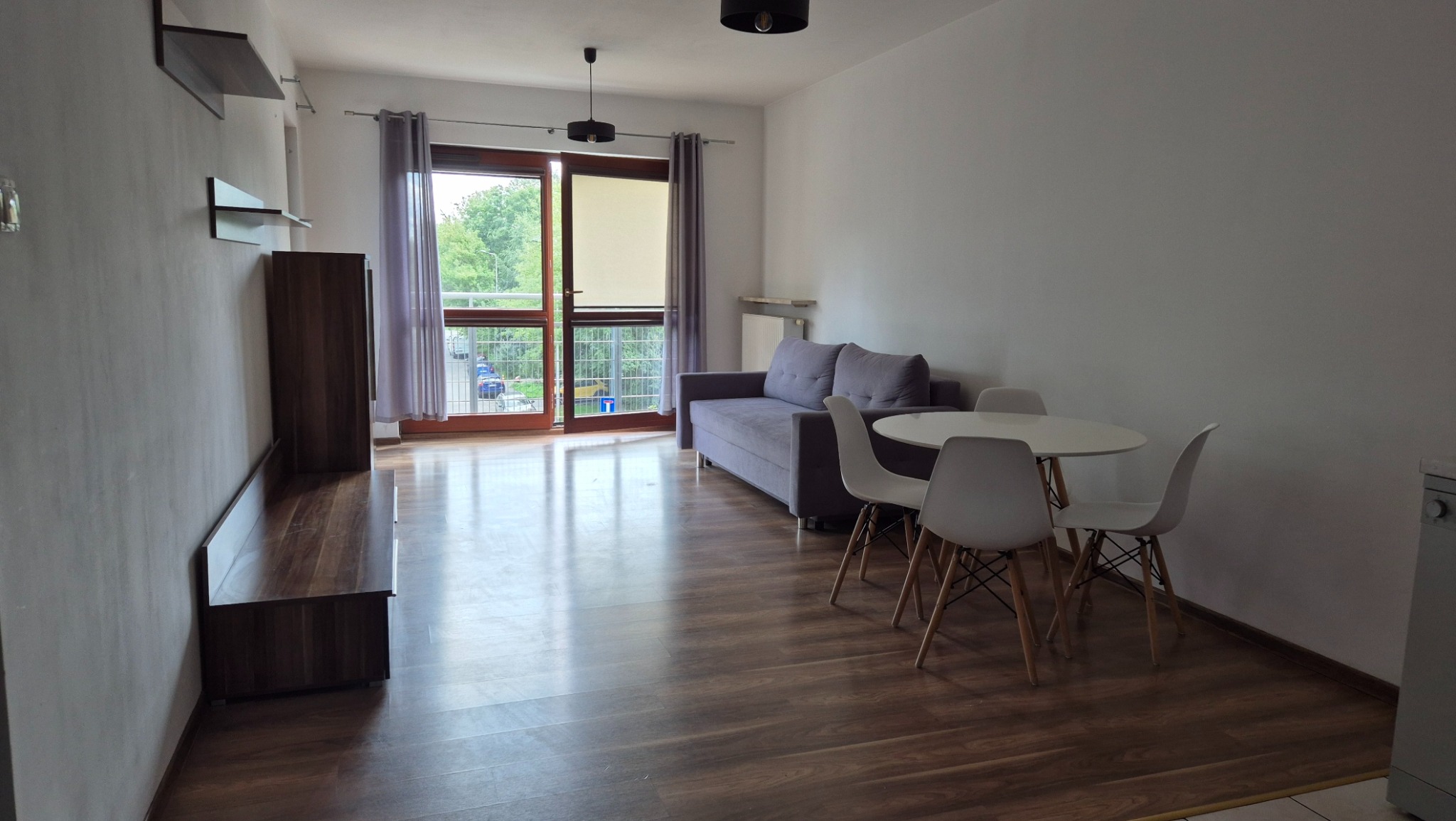 Jasne mieszkanie z balkonem: szara sofa, biały stół z krzesłami i drewniana podłoga. Minimalistyczny wystrój, widok na zieleń za oknem.