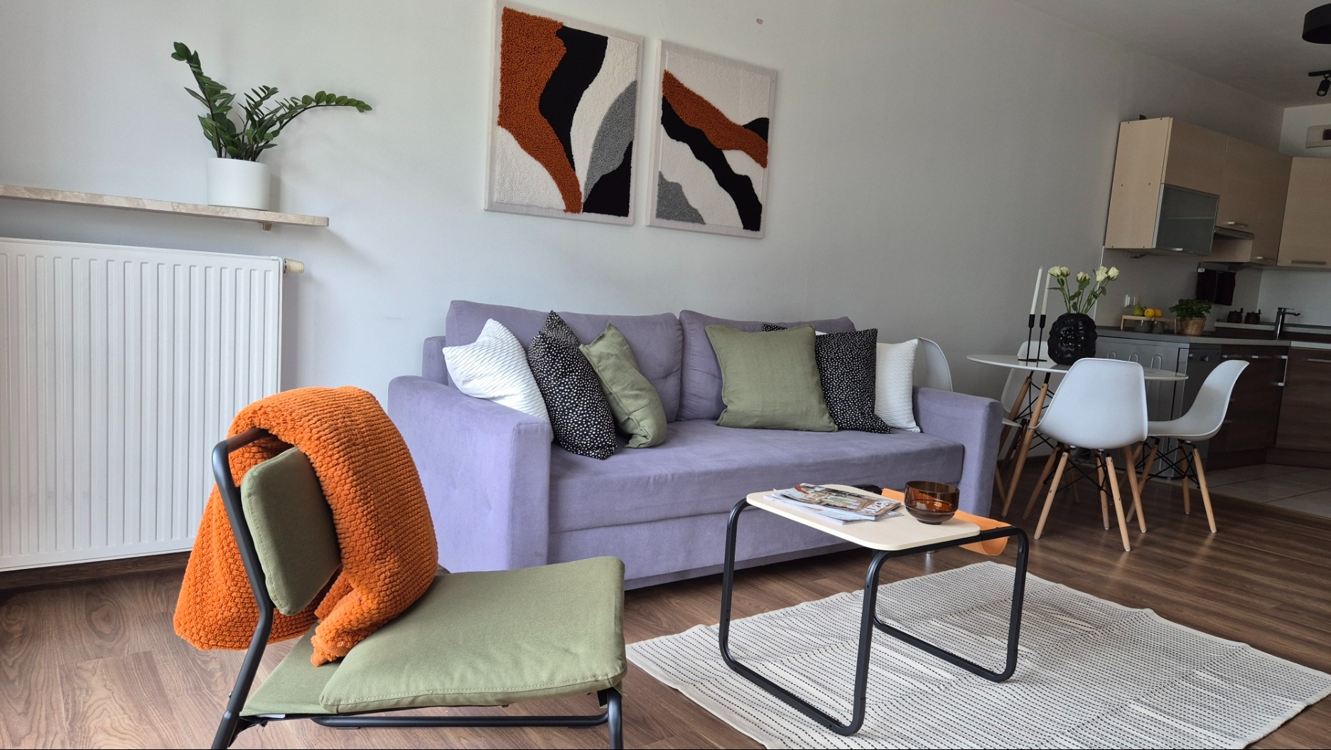 Jasny salon z fioletową sofą, zielonym fotelem i abstrakcyjnymi obrazami na ścianie. Na stole leżą gazety, a w tle widać aneks kuchenny.