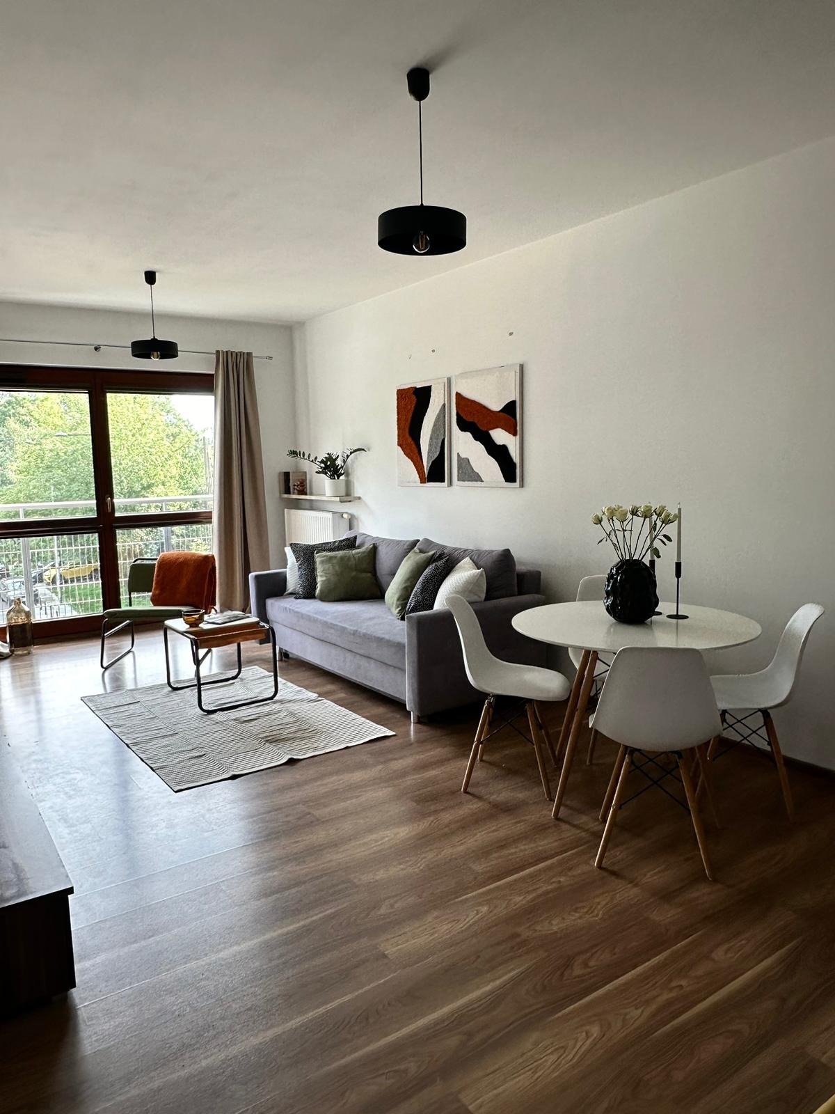 Jasne wnętrze mieszkania z drewnianą podłogą: szara sofa z poduszkami, okrągły stół z białymi krzesłami i abstrakcyjne obrazy na ścianie. Widok z balkonu na zieleń.