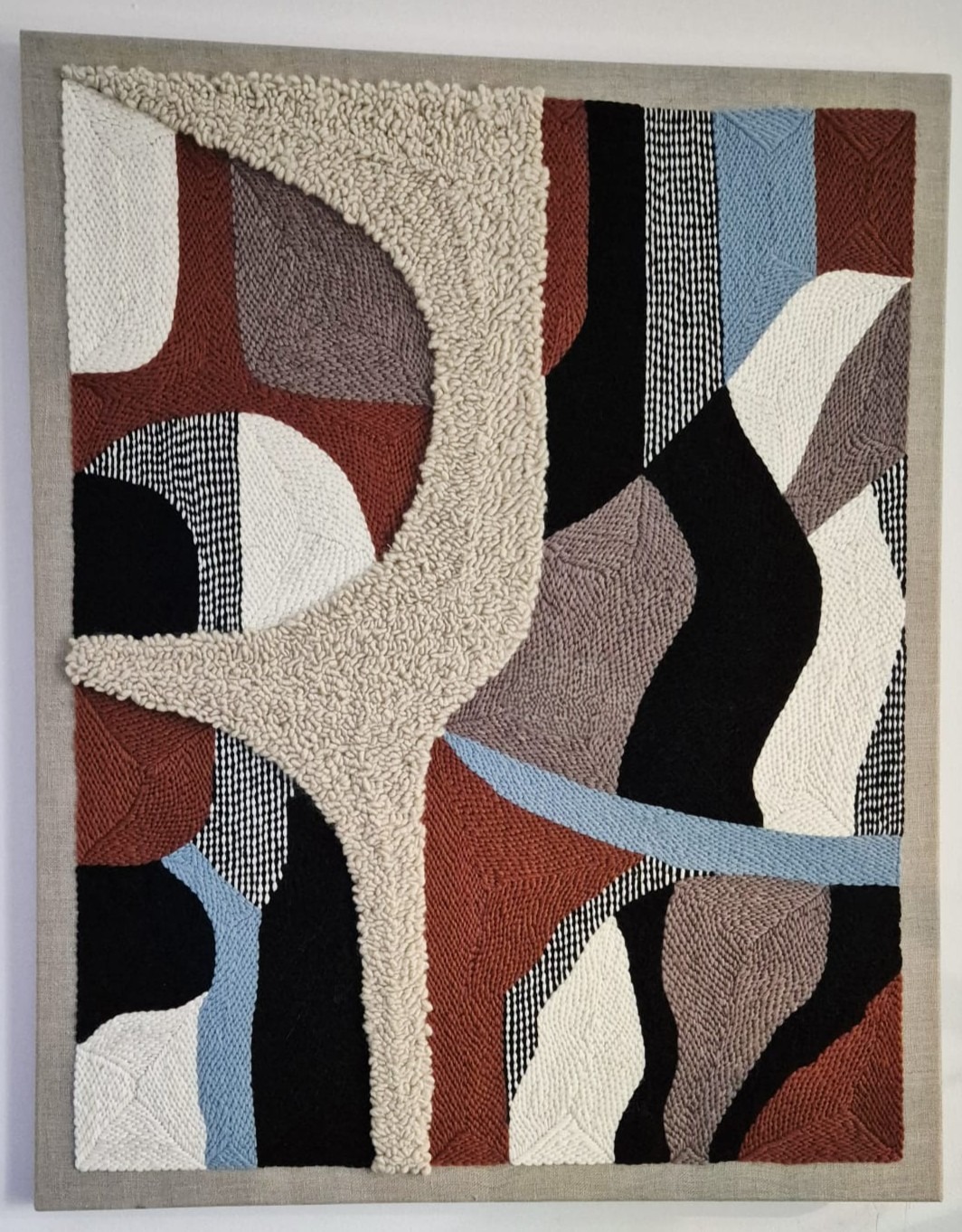 Haftowany obraz Punch Needle w geometrycznym stylu, z użyciem tekstur i kolorów: beż, brąz, czerń, biel i błękit. Dzieło rękodzielnicze w szarej ramie.