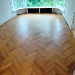 Parkiet ułożony w jodełkę w jasnym pokoju z oknem. Widoczne ślady po remoncie na ścianach i listwach przypodłogowych. Naturalne oświetlenie podkreśla wzór drewna.