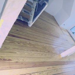 Parkiet Kamil - Ściana i sufit wykończone jasnym drewnem, widoczne okno i elementy konstrukcyjne zabezpieczone folią. Ukośne ujęcie podkreśla fakturę drewna i jasne wnętrze.