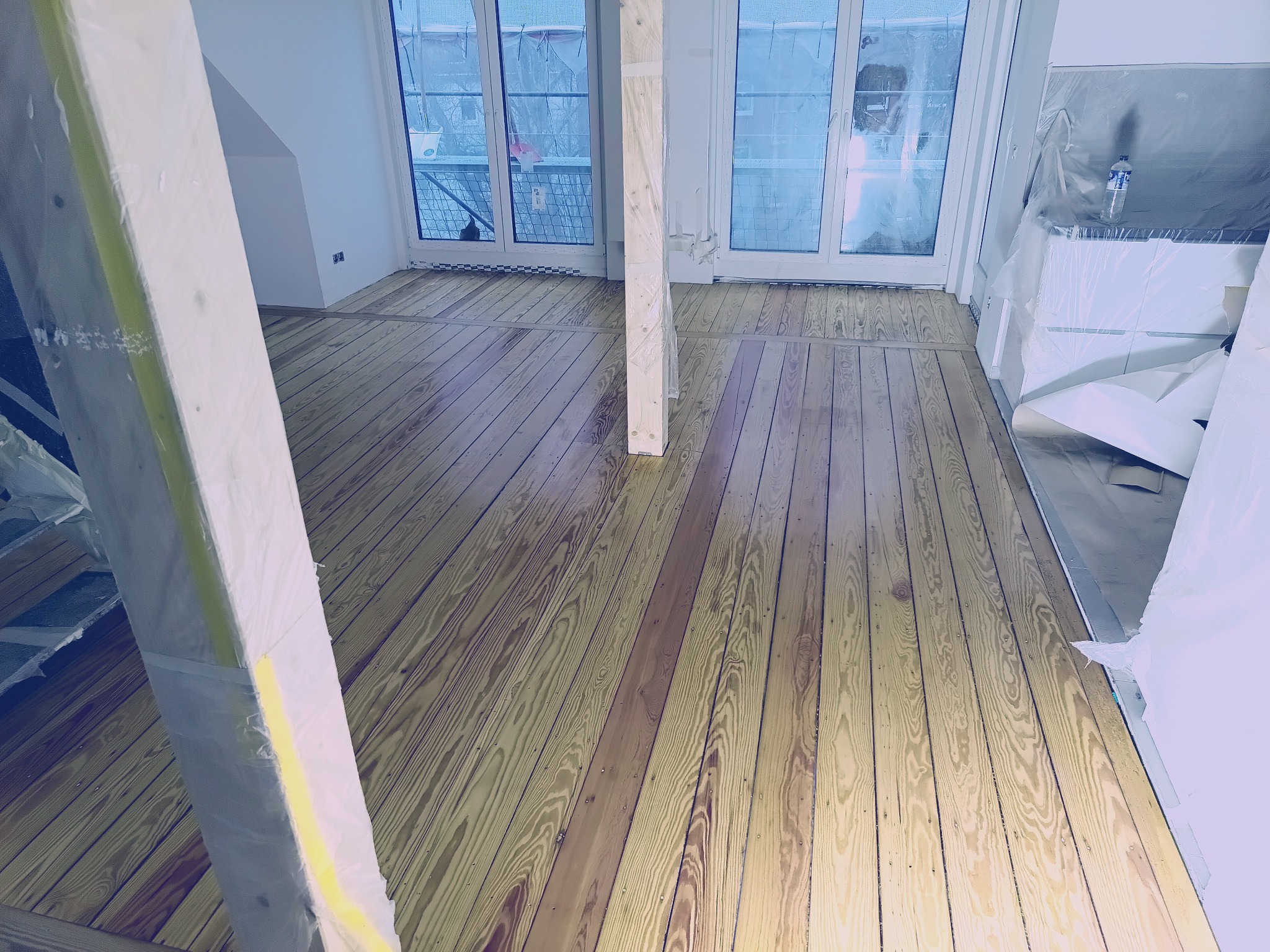 Jasne, drewniane deski podłogowe po renowacji, z widocznymi słupami zabezpieczonymi folią. Widok na przeszklony balkon w tle.