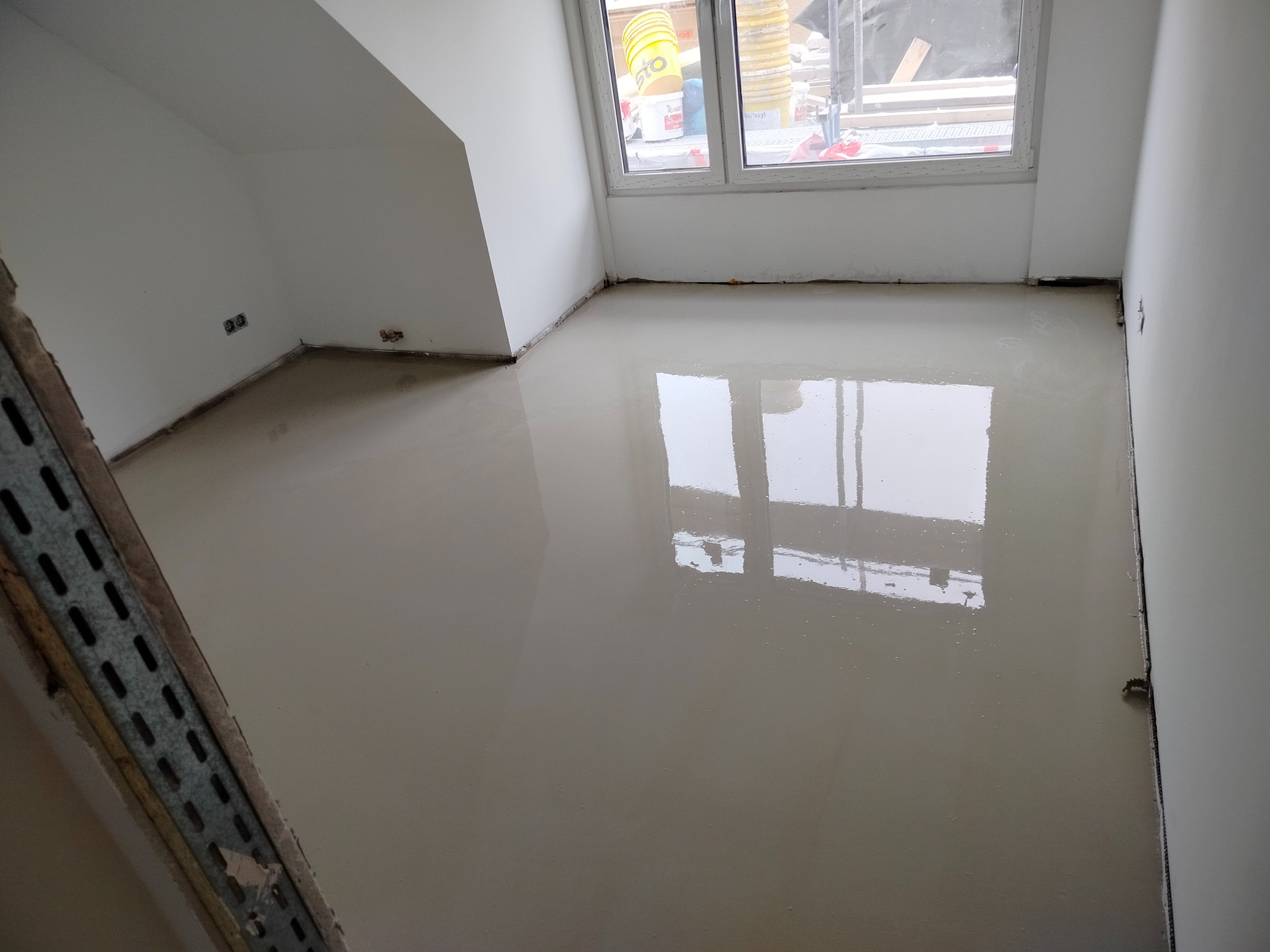 Wylana, gładka posadzka samopoziomująca w jasnym pokoju z oknem. Widoczne ślady po taśmie malarskiej przy ścianach. Metalowy profil przy ścianie po lewej.