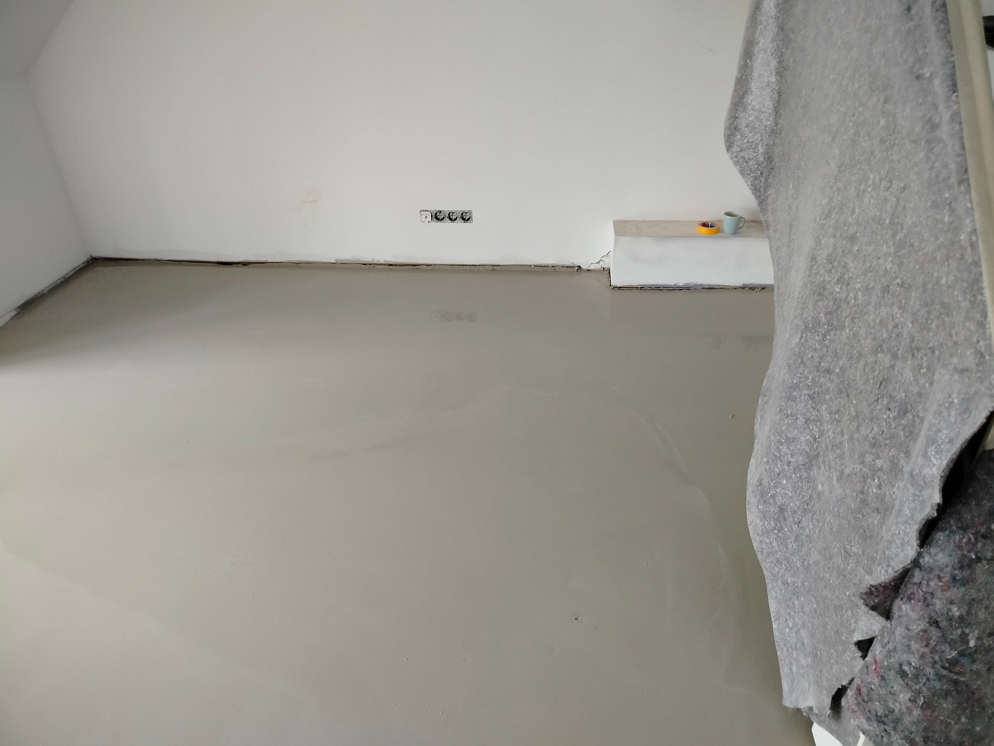 Wylany samopoziomujący podkład podłogowy w jasnym pomieszczeniu. Widoczne gniazdka elektryczne na ścianie i zabezpieczenie z folii malarskiej.