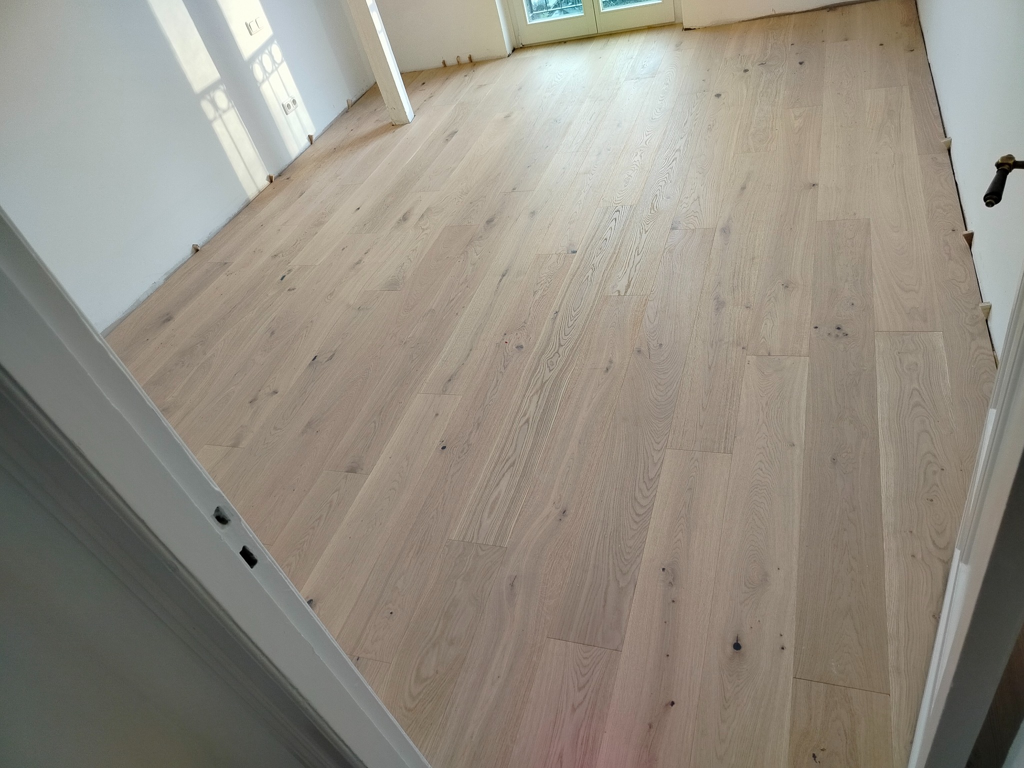 Jasna, drewniana podłoga z widocznymi słojami i sękami, świeżo ułożona w pustym pokoju z białymi ścianami i oknem w tle. Widoczne listwy przypodłogowe.