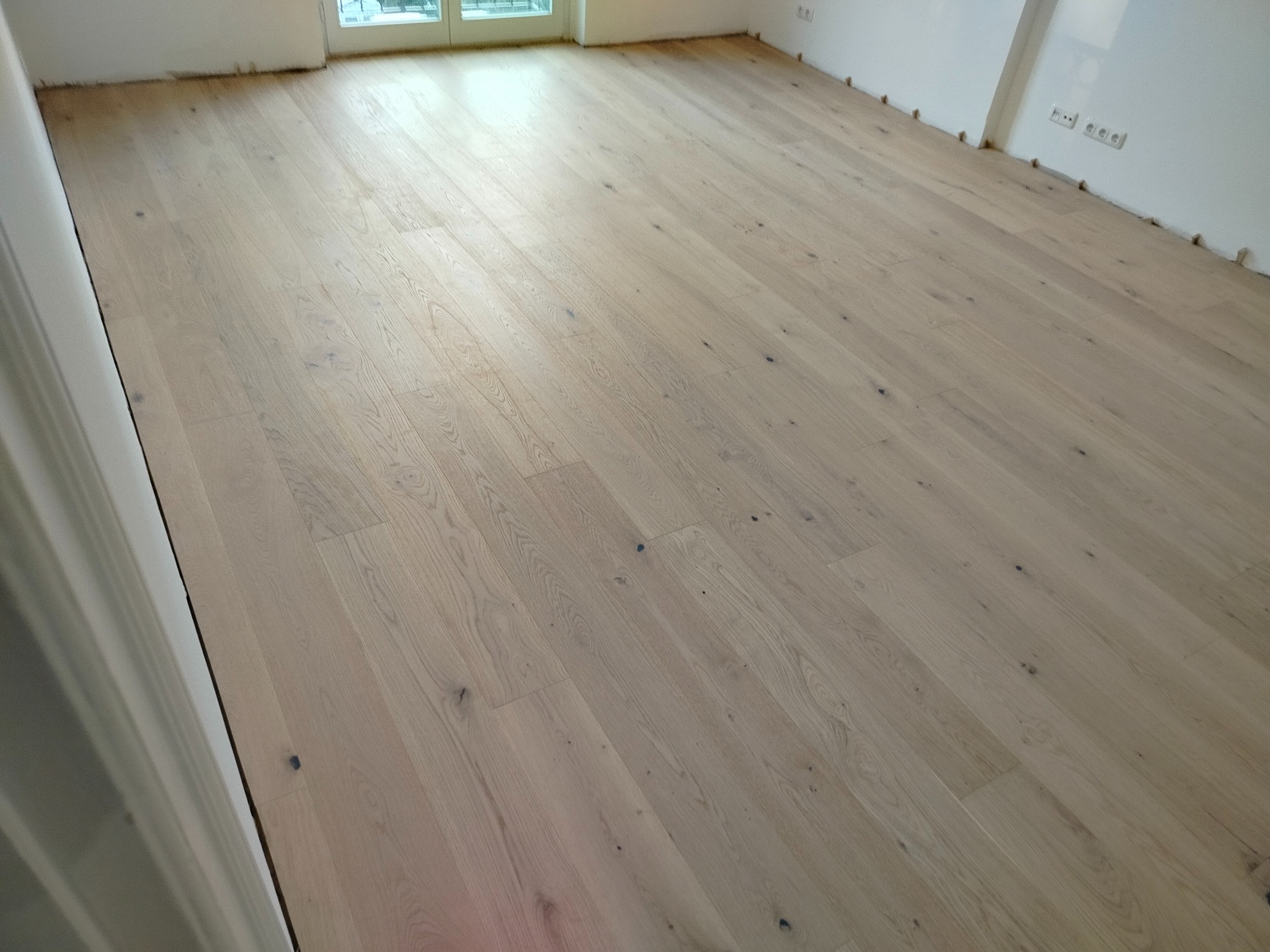 Jasny, drewniany parkiet ułożony w nowym mieszkaniu, widoczne listwy przypodłogowe i gniazdka elektryczne. Naturalne usłojenie drewna, ciepłe oświetlenie.