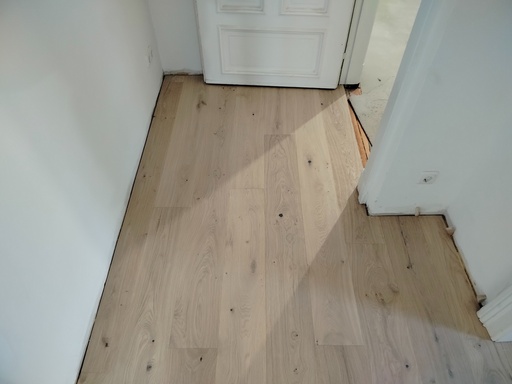 Jasne, drewniane panele podłogowe ułożone wzdłuż białej ściany i progu drzwi. Widoczne naturalne słoje drewna i subtelne niedoskonałości. Minimalistyczne wnętrze.