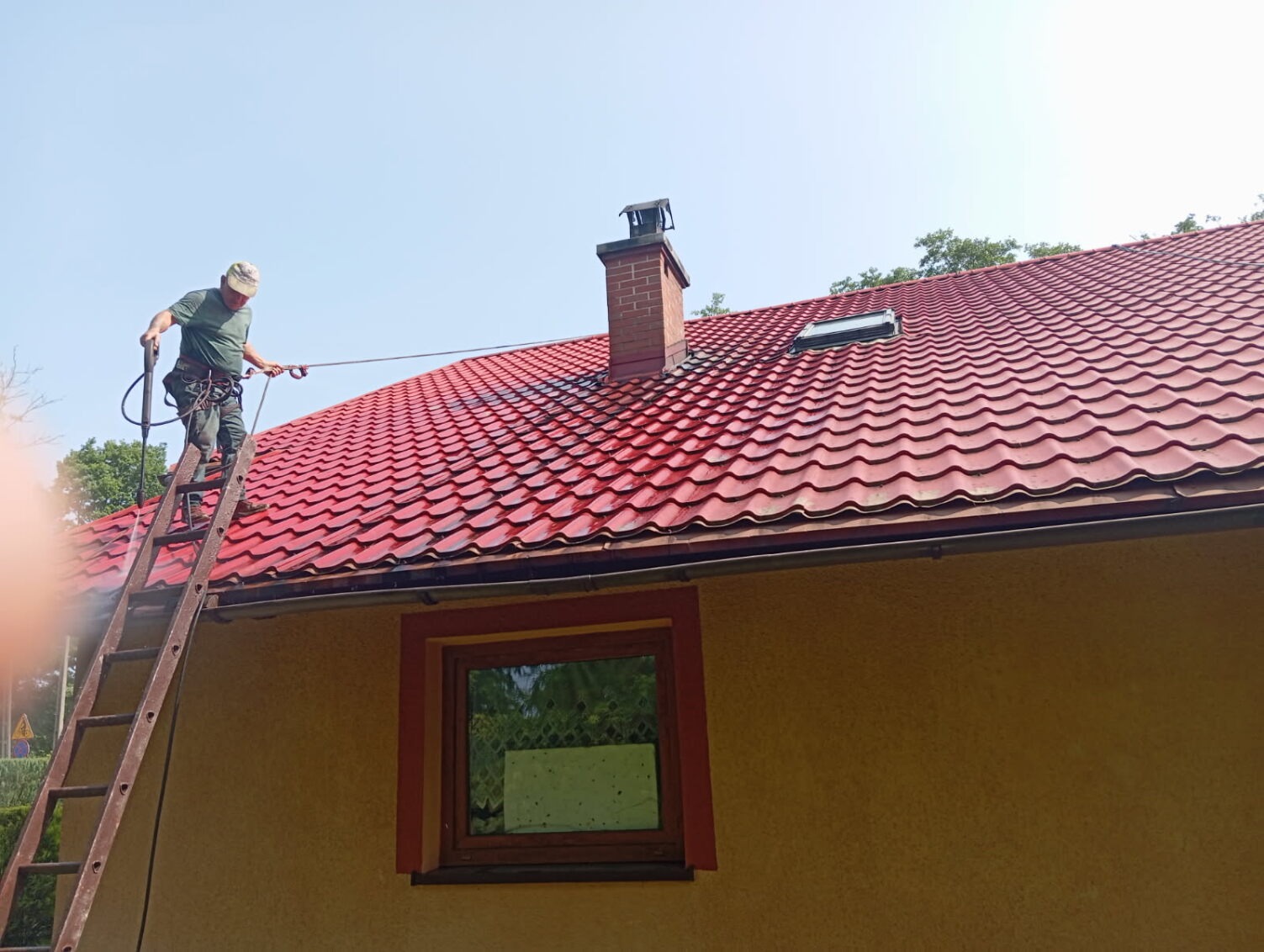 Pracownik na drabinie czyści czerwoną dachówkę myjką ciśnieniową. Widoczny komin i okno dachowe. Żółta elewacja budynku.