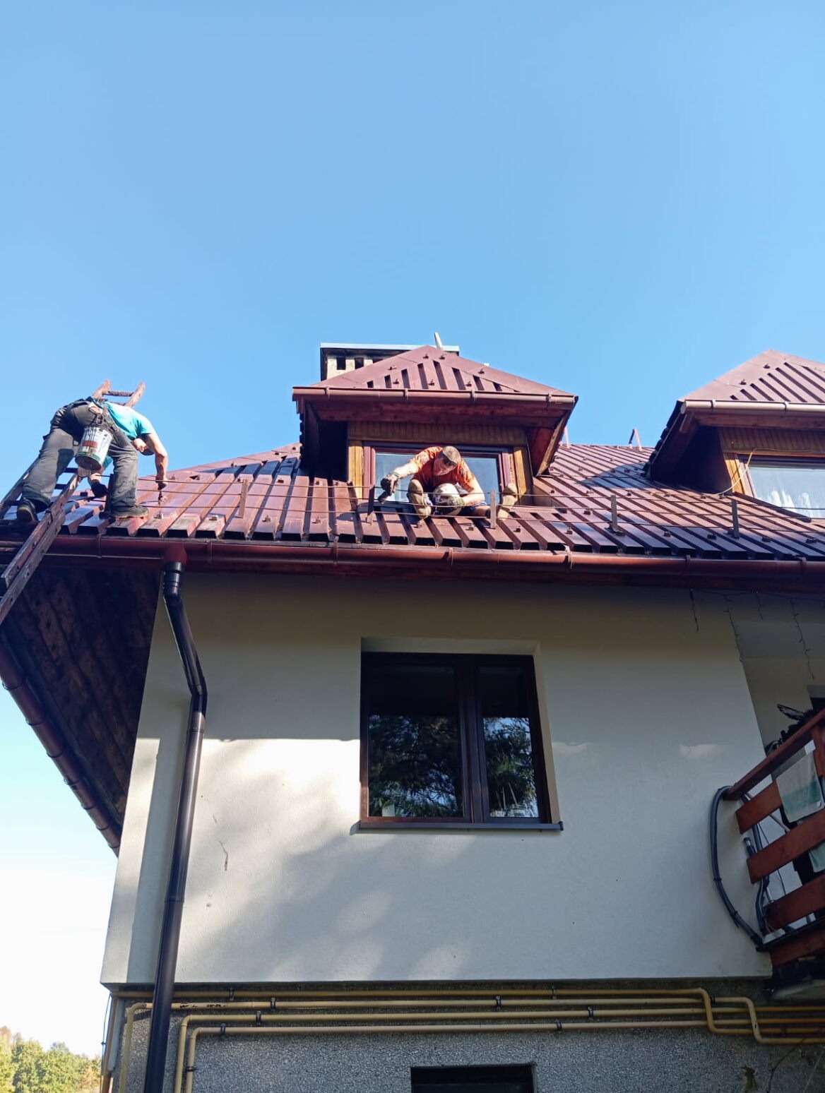 Dwóch dekarzy pracuje na dachu domu z brązową dachówką, jeden na drabinie, drugi przy oknie dachowym. Dach jest częściowo pokryty, widać rynny.