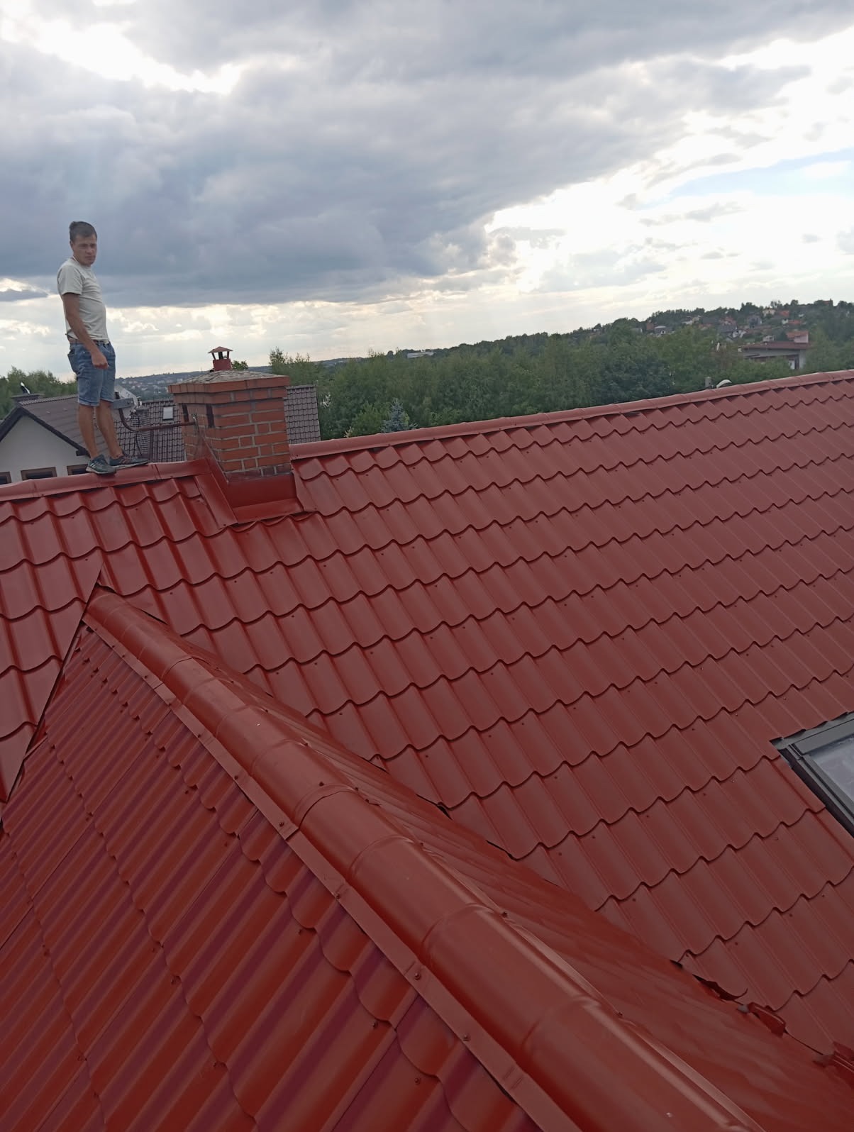 Czerwony dach z blachodachówki, widok z góry. Mężczyzna stoi na dachu obok komina z cegły. W tle zieleń i pochmurne niebo.