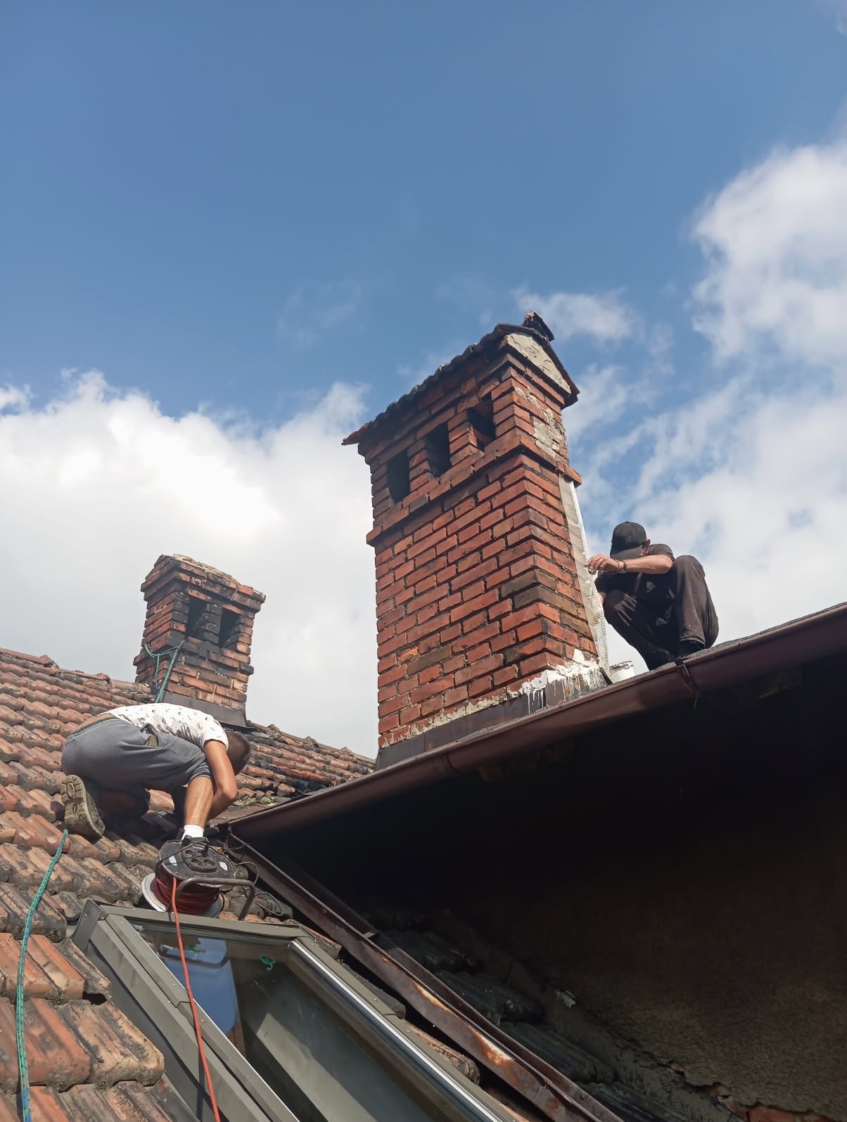 Prace na dachu: Dwóch mężczyzn zabezpiecza komin białą substancją. Widoczne ceglane kominy, dachówka i fragment okna dachowego na tle błękitnego nieba.