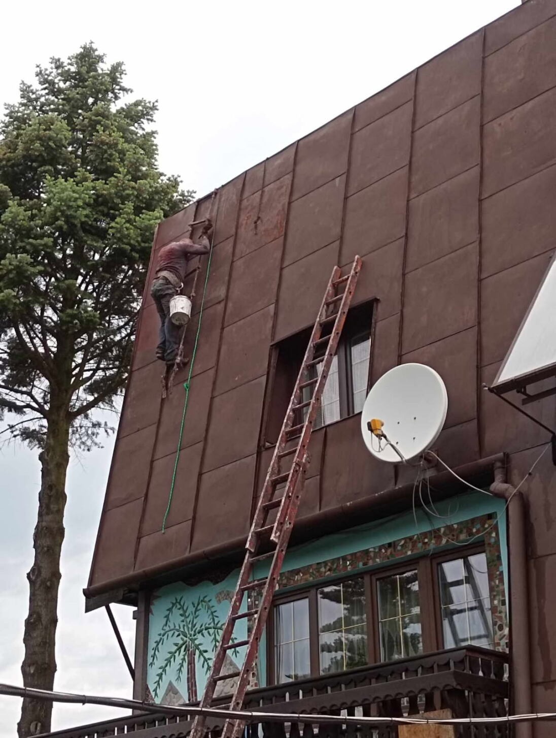 Pracownik na drabinie maluje brązowy, blaszany dach domu z anteną satelitarną. Widoczny fragment pomalowanej powierzchni i wiadro z farbą. Ujęcie z dołu.