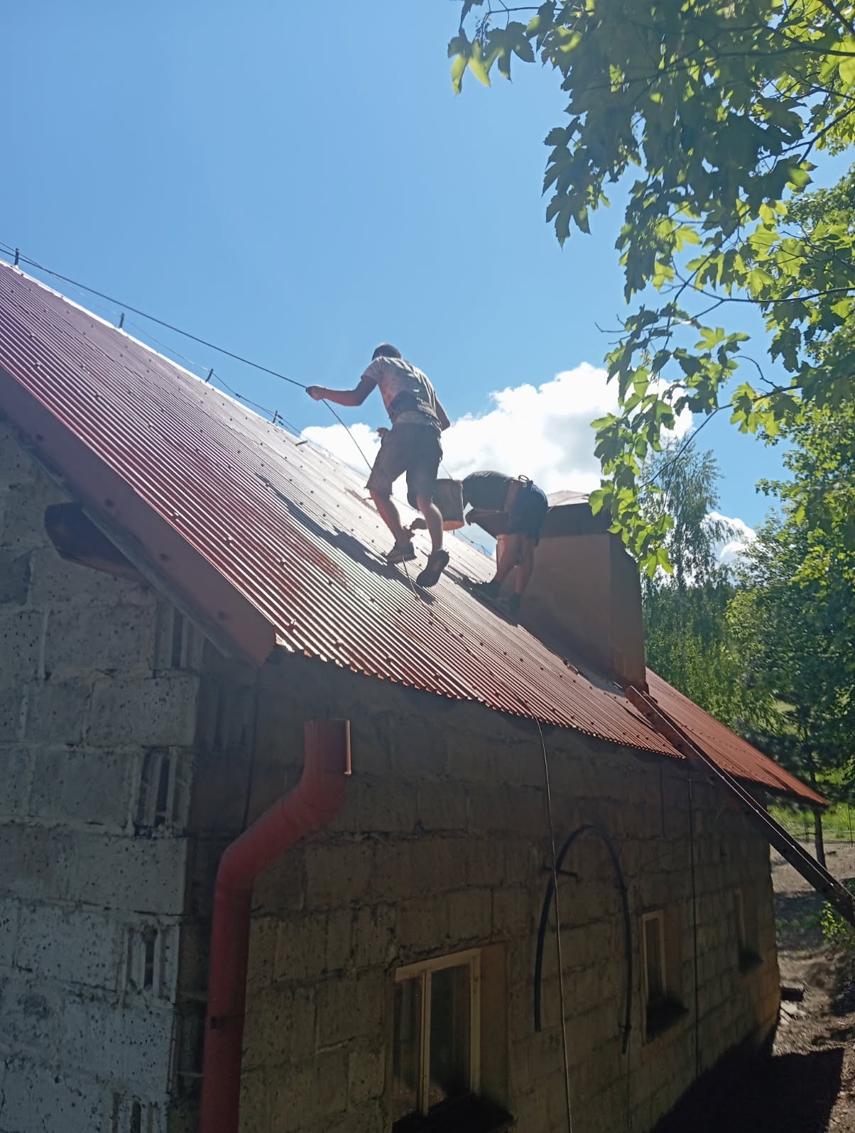 Dwóch dekarzy na czerwonym, falistym dachu domu z pustaków podczas pracy. Jeden z nich trzyma linę zabezpieczającą. Widok z dołu, w tle błękitne niebo i zieleń.