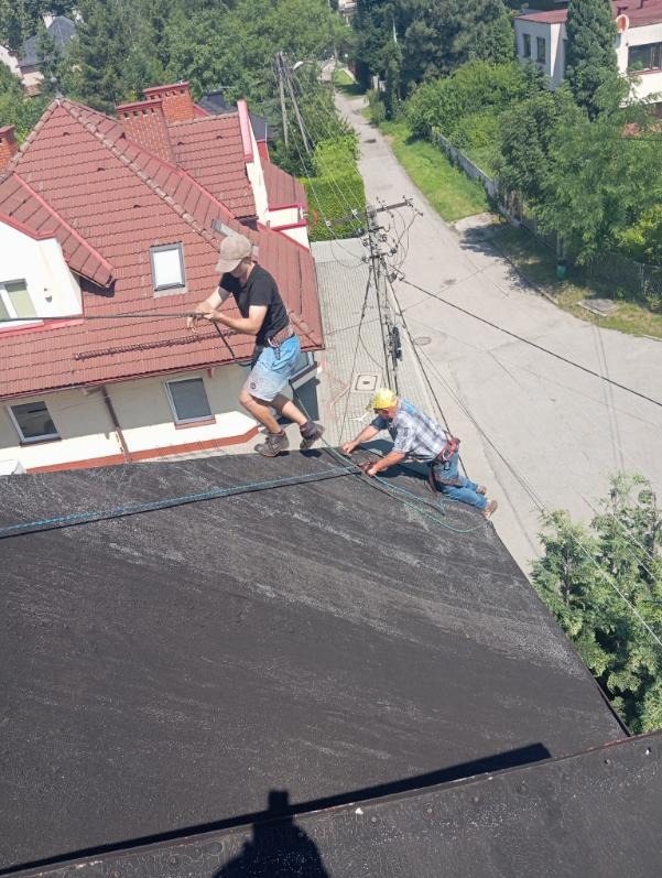 Dwóch dekarzy zabezpieczonych linami pracuje na płaskim, czarnym dachu. W tle budynki i zieleń. Ujęcie z góry, cień osoby robiącej zdjęcie na pierwszym planie.