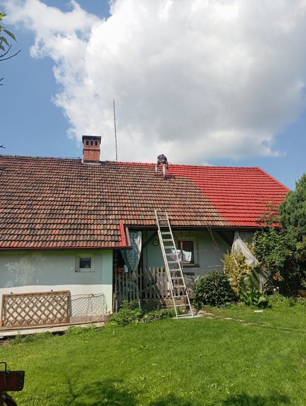Malowanie dachu na czerwono przez pracownika na drabinie. Połowa dachu pomalowana, druga w kolorze ceglastym. Widoczny komin i zielone otoczenie domu.