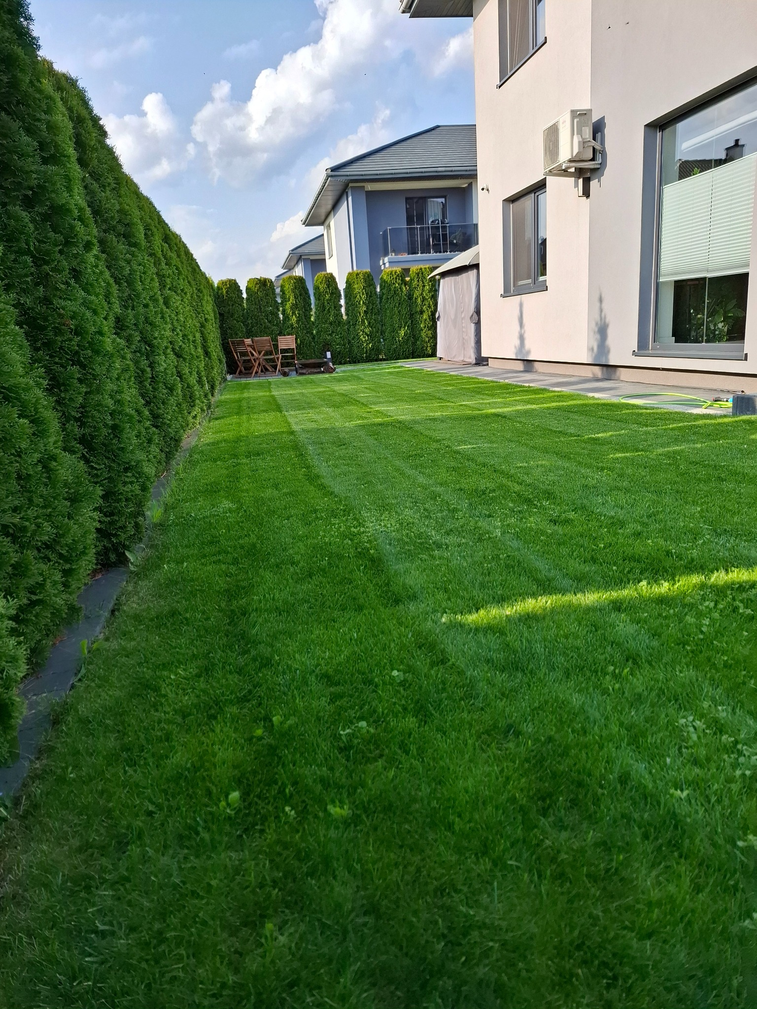 Perfekcyjnie skoszony trawnik w Skrzeszewie, z wyraźnymi pasami i zadbanymi tujami wzdłuż ogrodzenia. Widoczny fragment domu z klimatyzatorem.