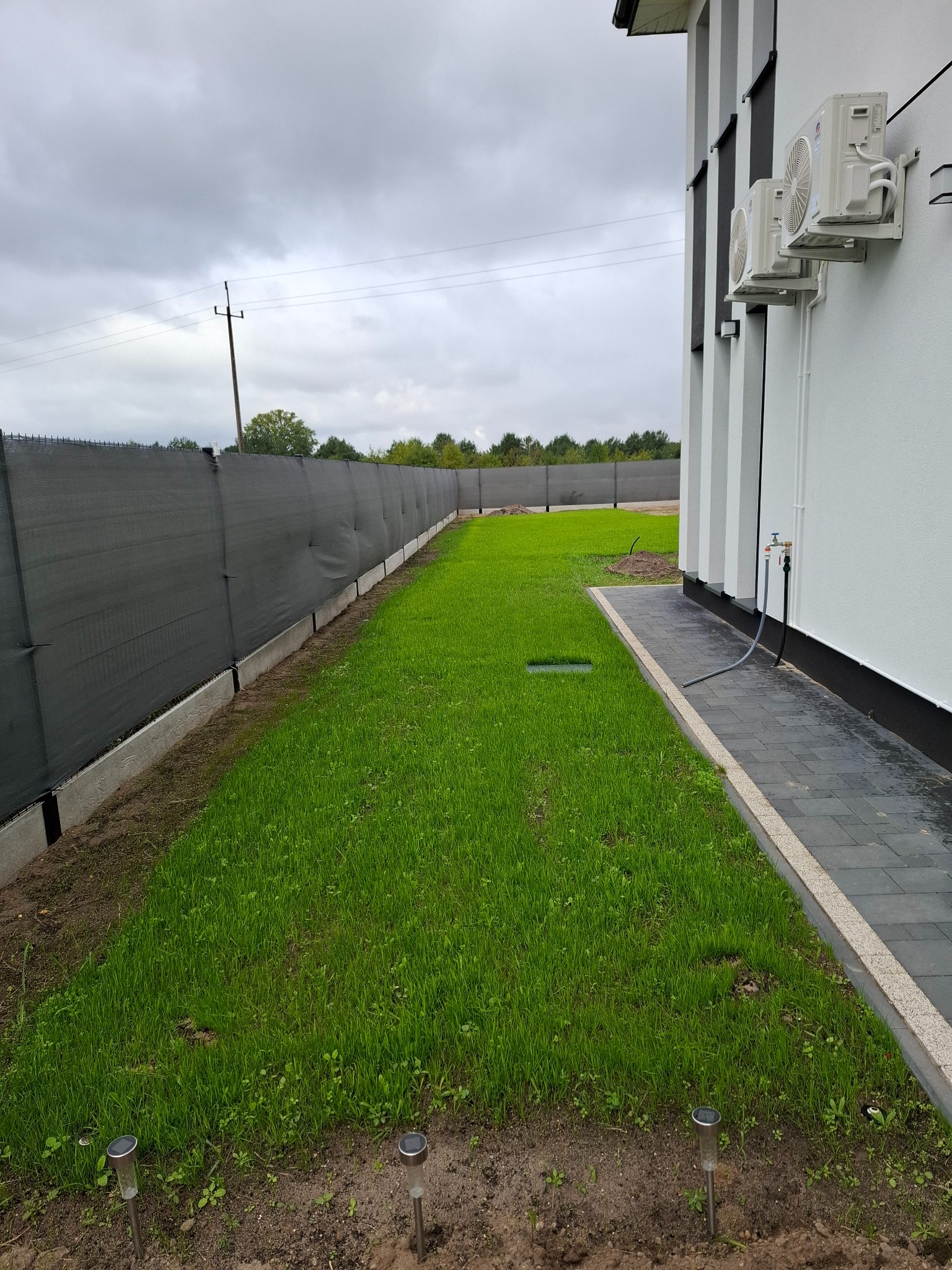 Nowo założony trawnik z widocznymi lampkami solarnymi, szarą siatką ogrodzeniową i fragmentem budynku z klimatyzacją. Świeża zieleń trawy po pracach.