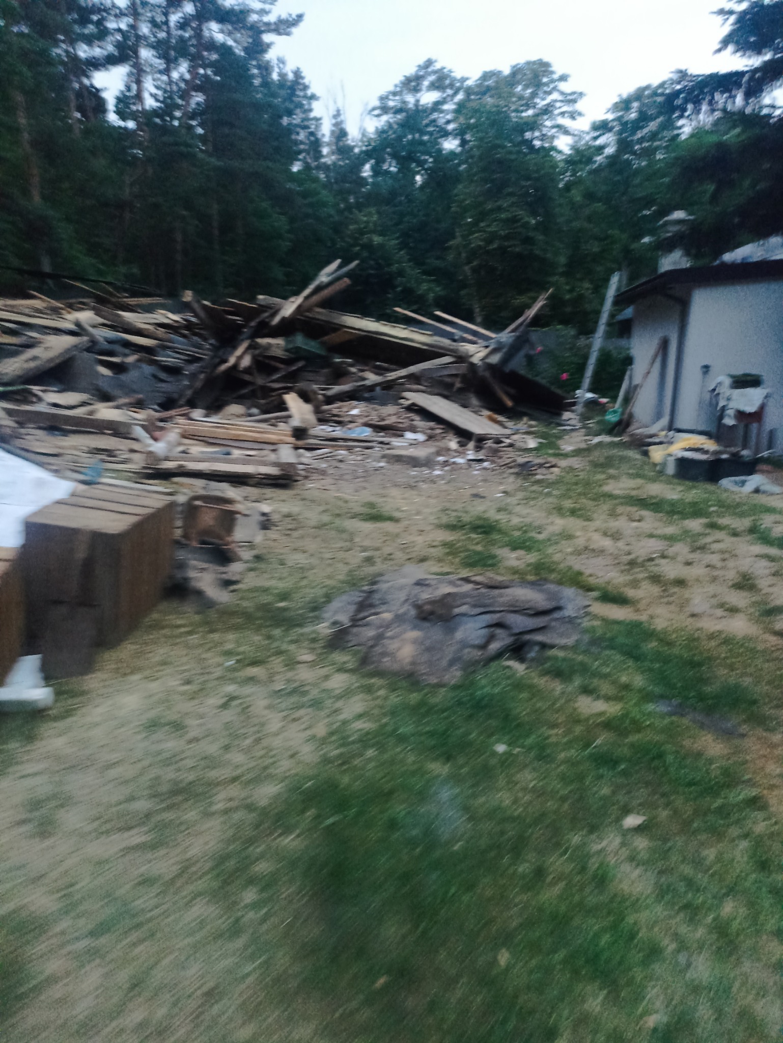 Stos drewnianych odpadów po rozbiórce budynku na zewnątrz, z fragmentem budynku i drzewami w tle. Widok na teren po zakończonych pracach rozbiórkowych.