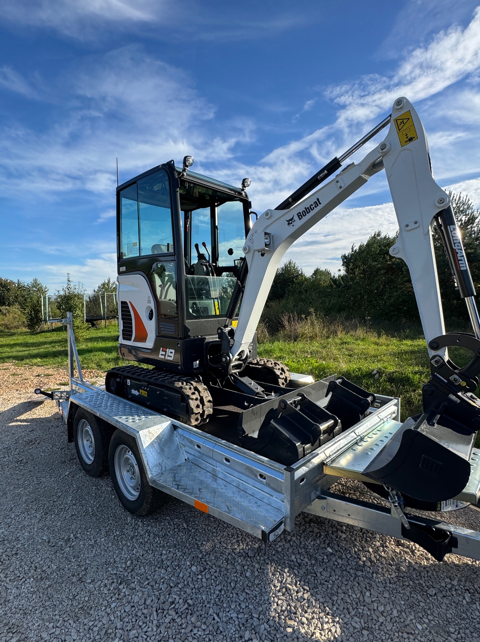 Kompaktowa minikoparka Bobcat E19 na dwuosiowej przyczepie, z dodatkowymi łyżkami. Błękitne niebo w tle. Widoczne detale konstrukcyjne i logo Bobcat.
