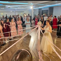 DJ DRS wesela eventy - Panna młoda w sukni ślubnej podczas oczepin, trzyma bukiet, otoczona gośćmi weselnymi z wstążkami. Zabawa weselna w sali z parkietem i dekoracjami.