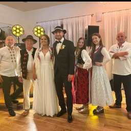 DJ DRS wesela eventy - Zespół weselny w strojach regionalnych, z nagłośnieniem i oświetleniem, pozuje z parą młodą w sali weselnej. Widoczny element dekoracji sali i parkiet.