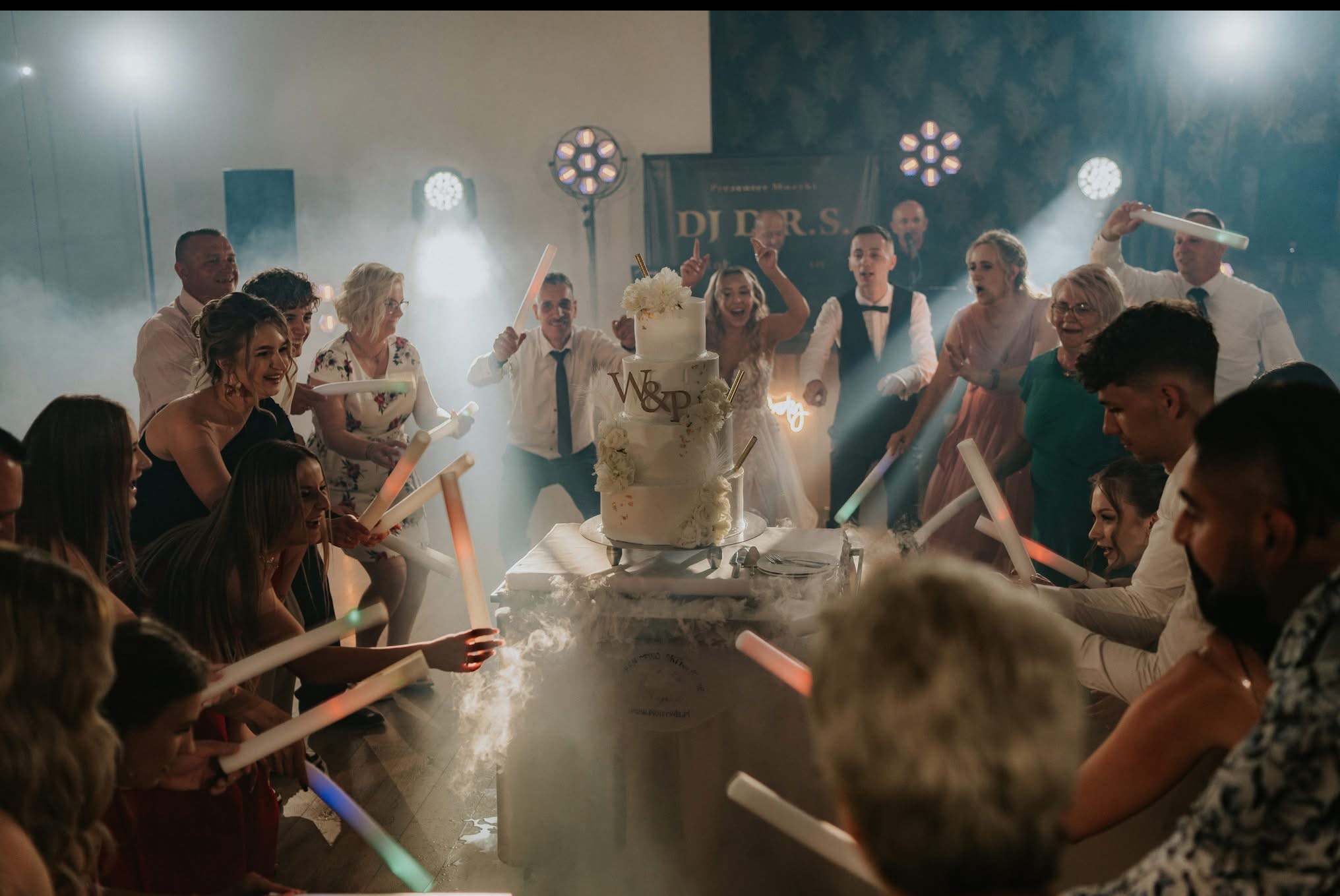 Goście weselni z podświetlanymi pałeczkami wokół tortu, w tle DJ i oświetlenie sceniczne. Efekt dymu dodaje magii wydarzeniu.
