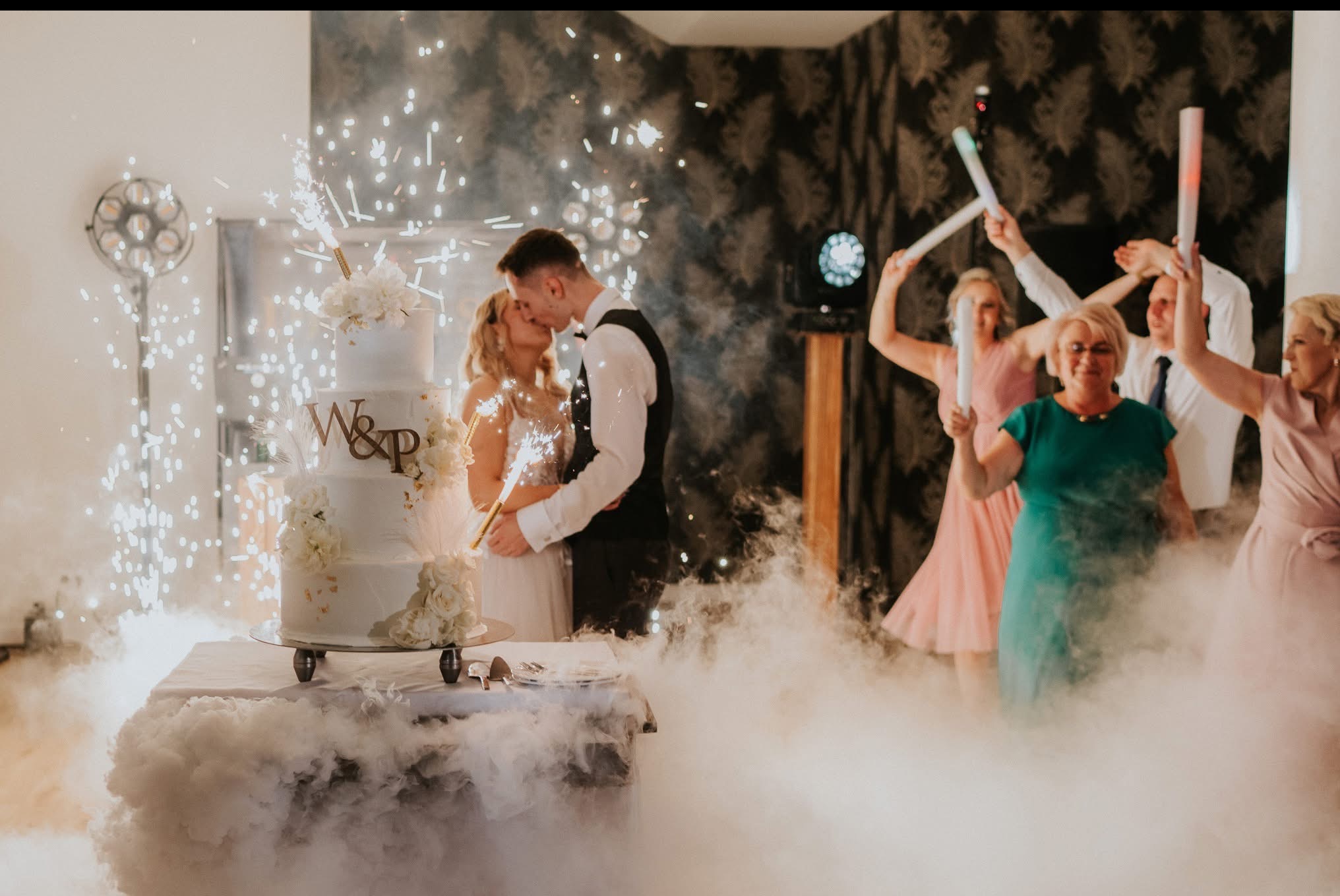 Para młoda kroi tort weselny z fontannami iskier, w tle goście z ledowymi pałeczkami, dekoracja sali z efektem ciężkiego dymu.
