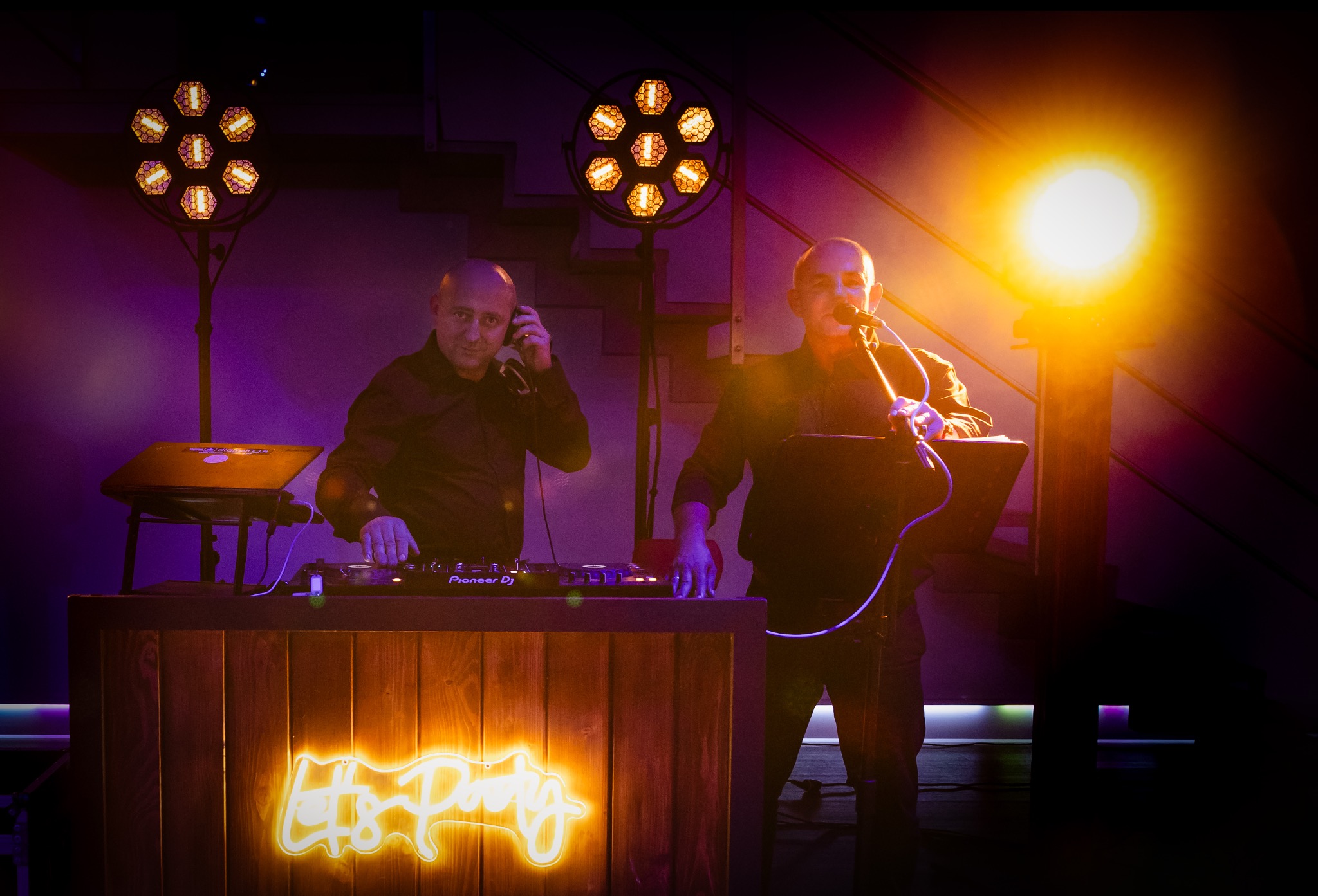 Dwóch DJ-ów za drewnianym stanowiskiem z neonem 'Let's Party', oświetleni reflektorami. Jeden w słuchawkach, drugi z mikrofonem. Scena w fioletowych barwach.