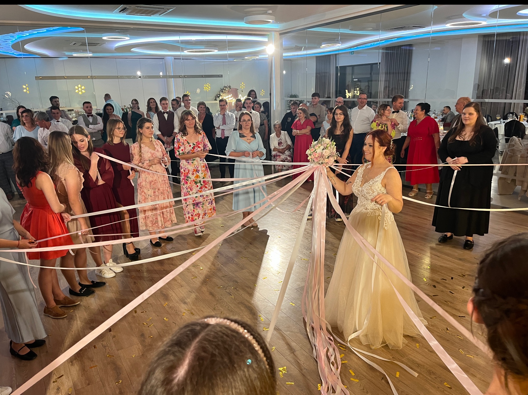 Panna młoda w sukni ślubnej podczas oczepin, trzyma bukiet, otoczona gośćmi weselnymi z wstążkami. Zabawa weselna w sali z parkietem i dekoracjami.