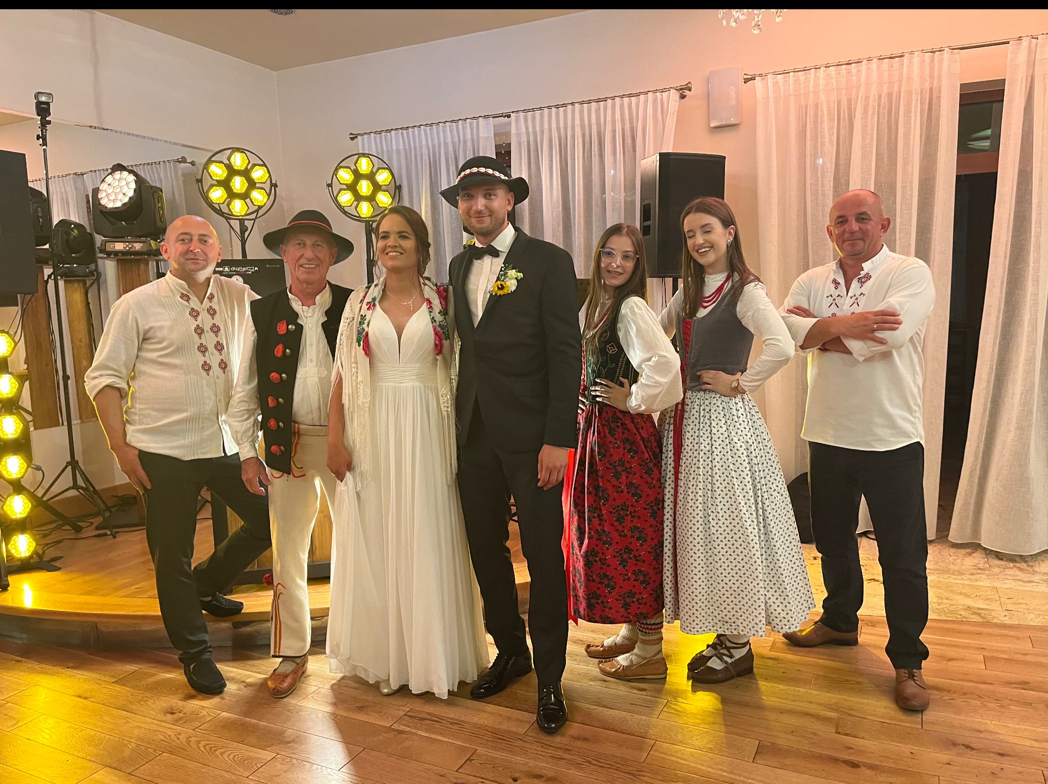Zespół weselny w strojach regionalnych, z nagłośnieniem i oświetleniem, pozuje z parą młodą w sali weselnej. Widoczny element dekoracji sali i parkiet.
