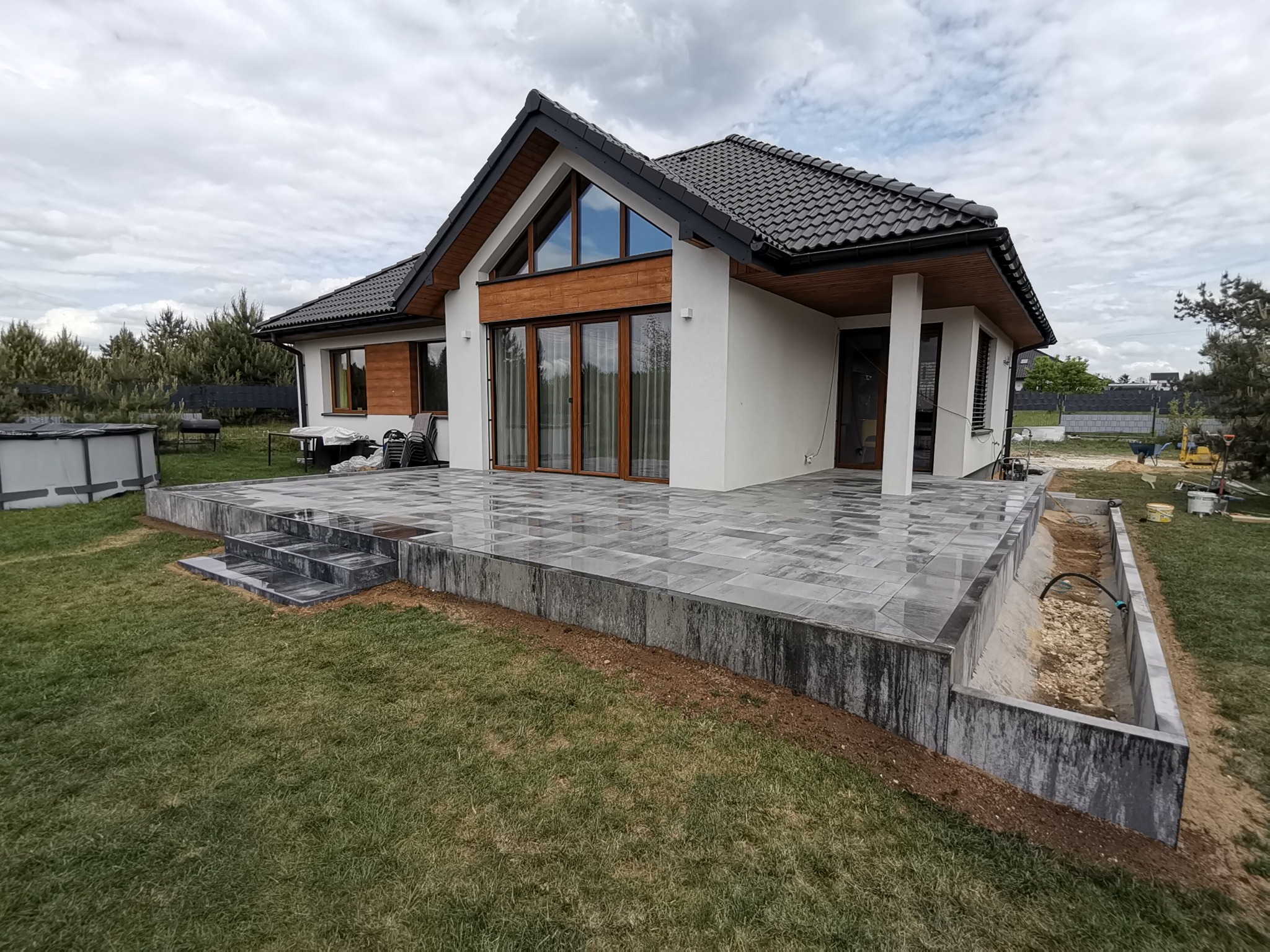 Nowoczesny dom z tarasem wyłożonym szarą kostką brukową. Widoczne schody z kostki, trawnik i fragment systemu odwadniającego. Pochmurne niebo w tle.
