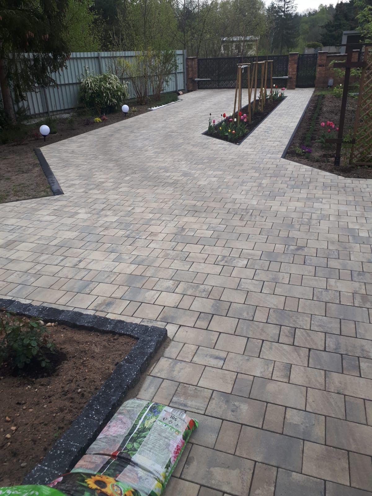 Jasnoszara kostka brukowa tworzy wzór na podjeździe z rabatami kwiatowymi. W tle ogrodzenie z metalu i siatki.
