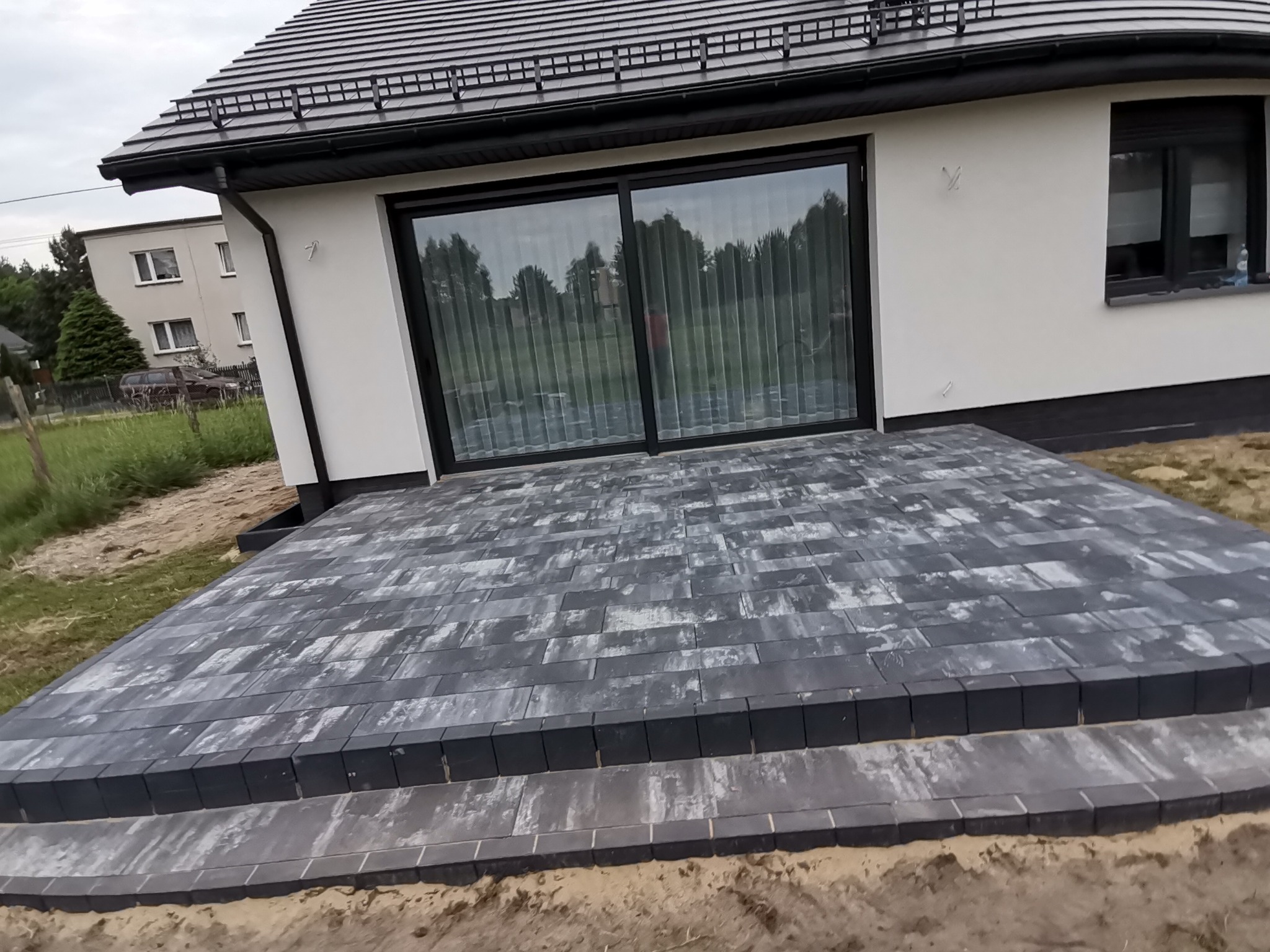 Nowoczesny taras z szarej kostki brukowej przy domu z dużym oknem przesuwnym. Kostka ułożona w regularny wzór, widoczne obrzeża i przygotowane podłoże.