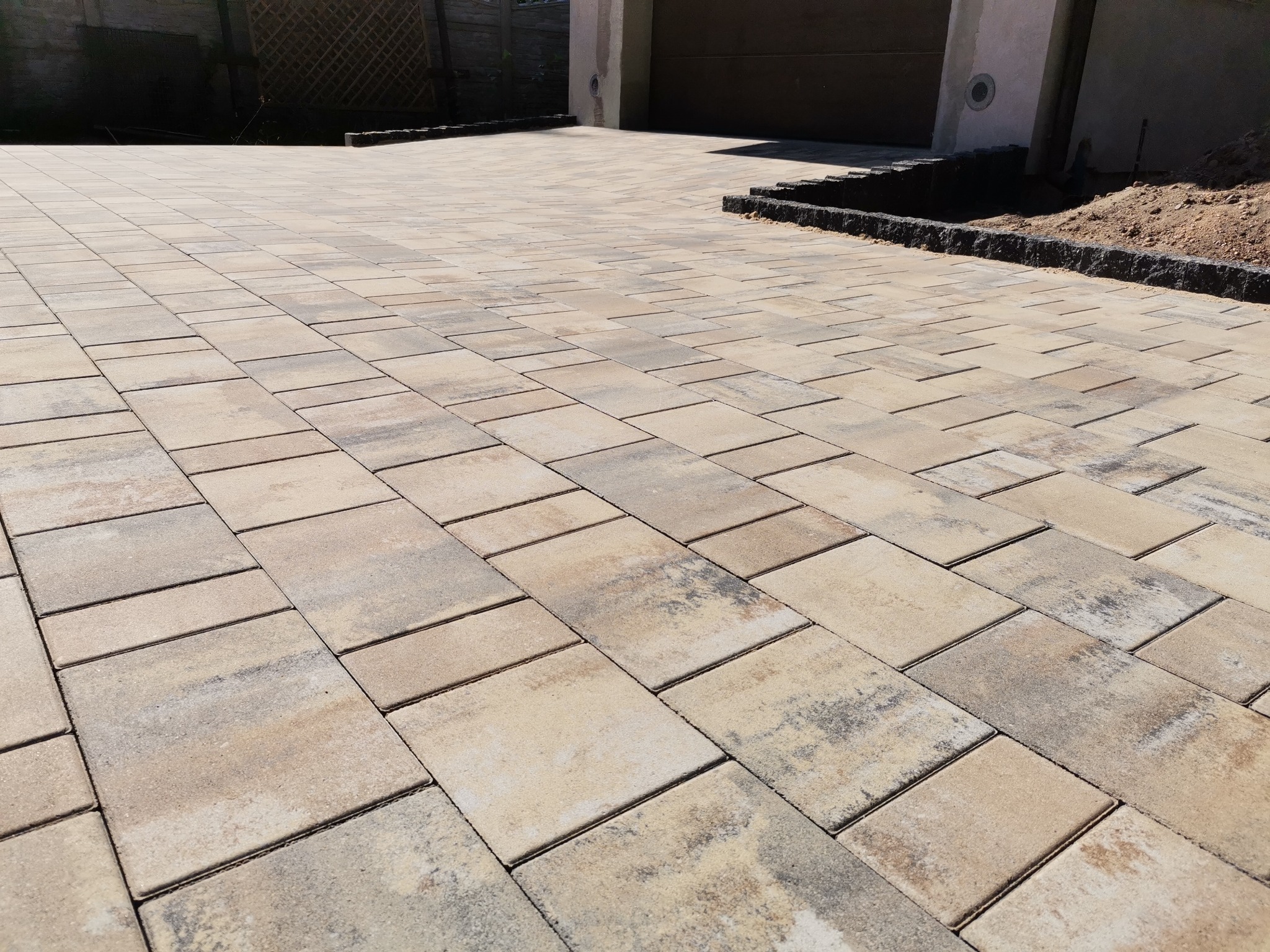 Jasna, beżowo-szara kostka brukowa ułożona na podjeździe, obrzeża z ciemnego kamienia, w tle brama garażowa i fragment ogrodzenia, słoneczny dzień.