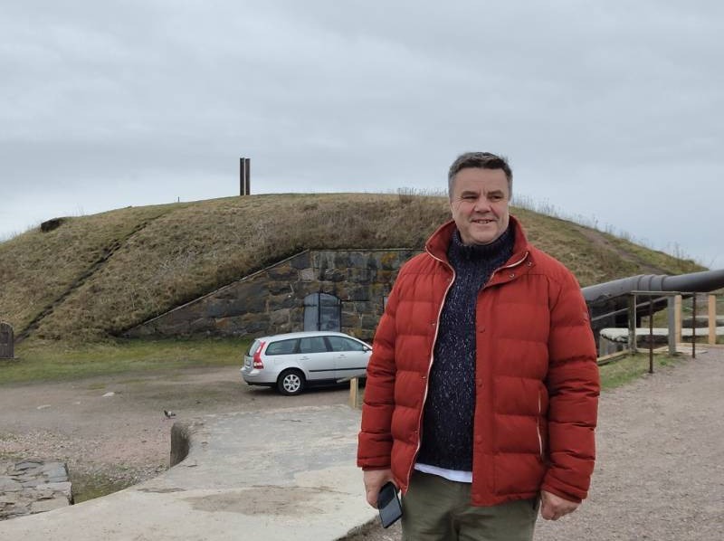 Mężczyzna w czerwonej kurtce na tle trawnika z ukrytym wejściem i armatą, pochmurne niebo. Nietypowe ujęcie krajobrazu z elementami architektury ziemnej.