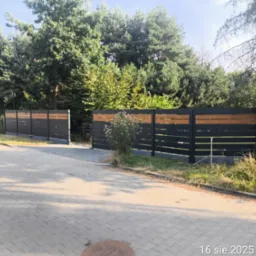 Nowoczesne ogrodzenie posesji z czarnych paneli z drewnianymi akcentami, widok z zewnątrz na tle zieleni. Tablica ograniczenia prędkości w tle.