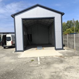 Tomasz Mech - Szara, metalowa hala z otwartymi wrotami, widoczna drabina wewnątrz. Na zewnątrz zaparkowany biały bus, obok betonowe ogrodzenie. Błękitne niebo w tle.