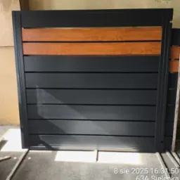 Nowoczesna, przesuwna furtka z szarych, metalowych paneli i drewnianych akcentów. Widoczne detale konstrukcyjne i elementy mocujące. Furtka w trakcie montażu.