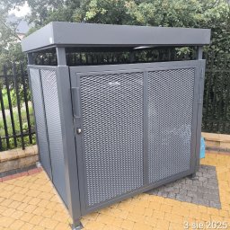 Tomasz Mech - Szara, metalowa obudowa na śmietniki z ażurowymi drzwiami, na kostce brukowej. W tle czarne ogrodzenie i zieleń.