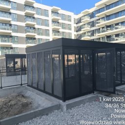 Tomasz Mech - Nowoczesna, ażurowa konstrukcja metalowa w kolorze czarnym, prawdopodobnie na balkonie lub tarasie, z budynkiem w tle. Otwarta brama z siatki.