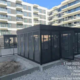 Nowoczesna, ażurowa konstrukcja metalowa w kolorze czarnym, prawdopodobnie na balkonie lub tarasie, z budynkiem w tle. Otwarta brama z siatki.