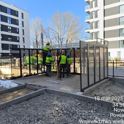 Tomasz Mech - Montaż ażurowej konstrukcji przy nowym budynku. Pracownicy w odblaskowych kamizelkach. Widoczne elementy konstrukcyjne i narzędzia. Słoneczny dzień.