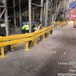 Tomasz Mech - Żółta barierka ochronna z metalu w hali przemysłowej, pracownicy w kaskach wykonują prace montażowe. Widoczne elementy konstrukcyjne i wózek na śmieci.