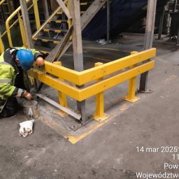 Tomasz Mech - Pracownik w kasku maluje żółtą farbą metalową konstrukcję ochronną w hali przemysłowej. Widoczne schody, podłoga z zabrudzeniami i datownik.
